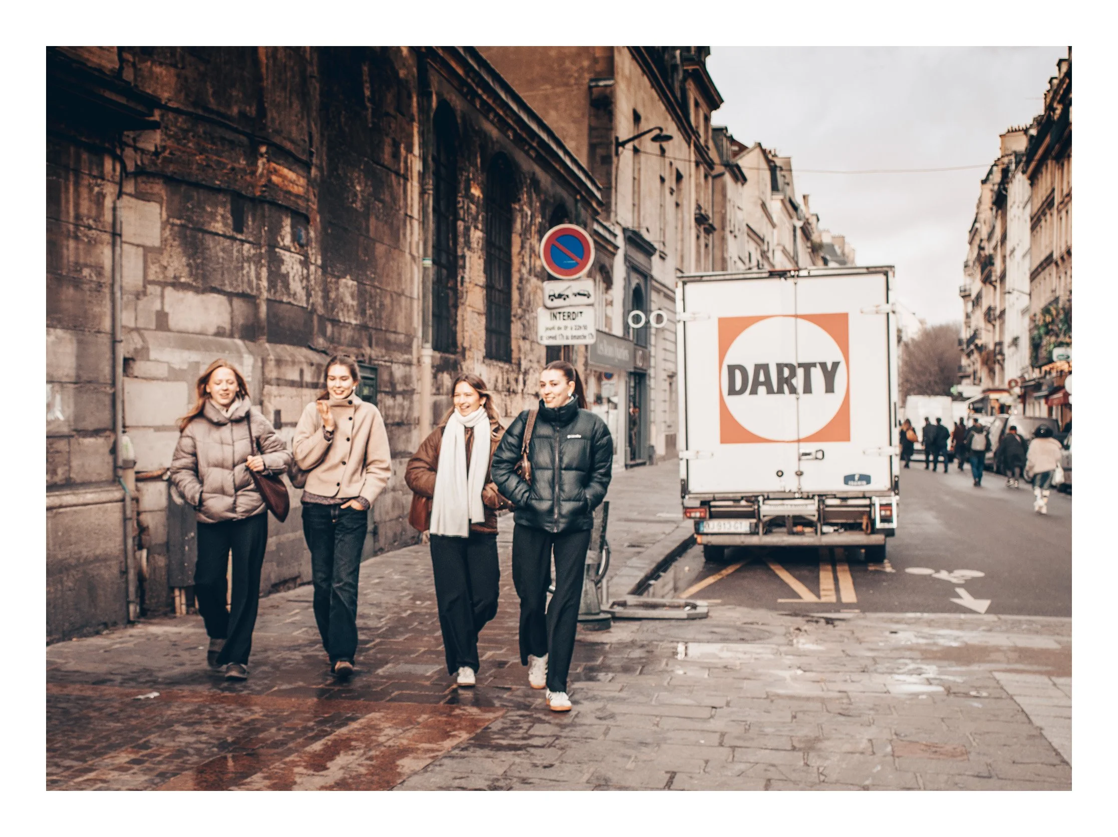 Four women walking on a city street, smiling and chatting, with historical buildings and a delivery truck with 'DARTY' logo in the background.