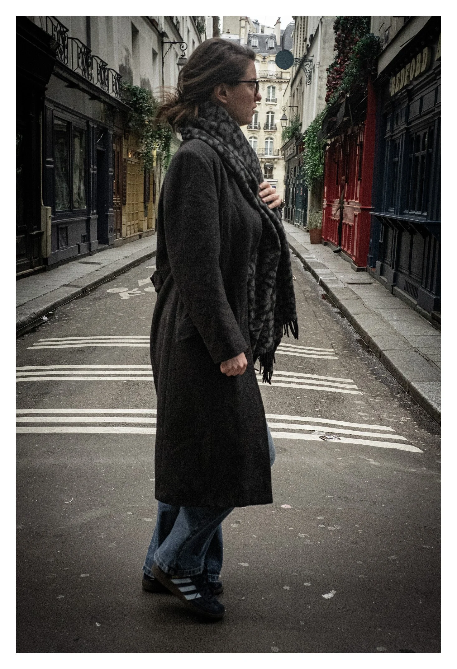 A woman standing on a city street corner, dressed in a long black coat, jeans, and Adidas sneakers, with a scarf around her neck, in an urban area with colorful buildings and flower decorations.