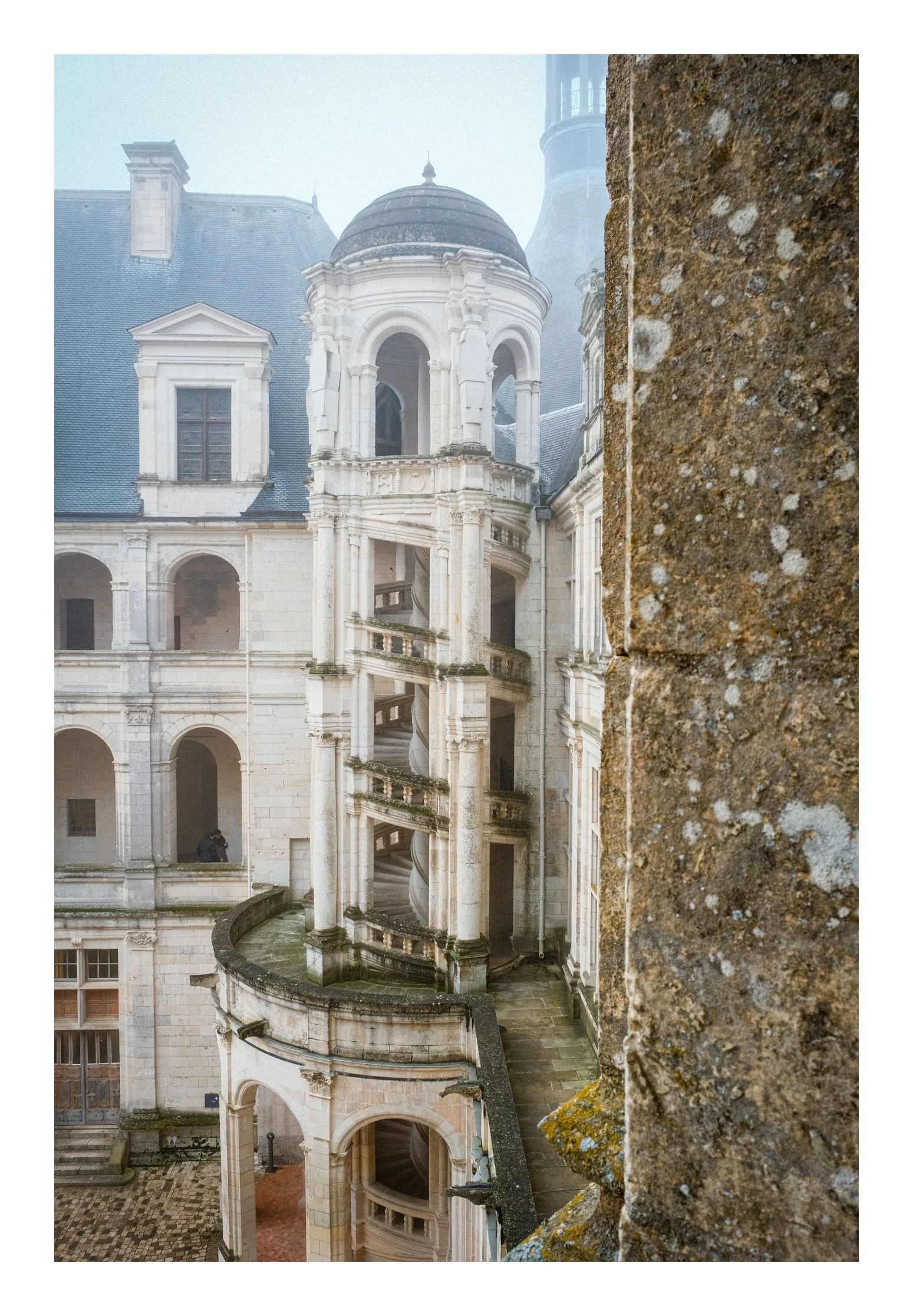 View of an old European castle or palace with a spiral staircase visible through a window, foggy weather, and a stone wall in the foreground.