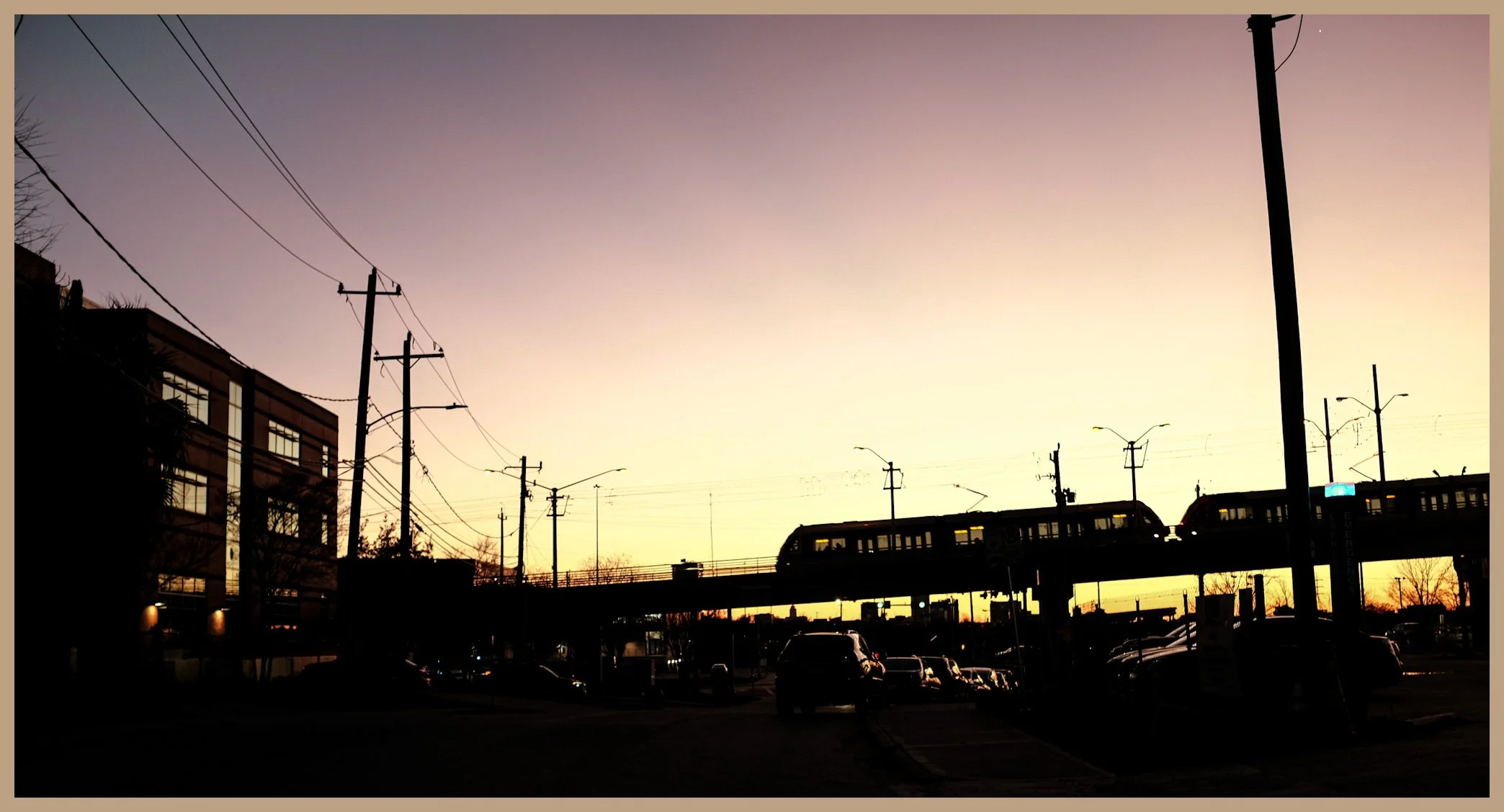Silhouette of a cityscape at sunset with electric poles, a moving train on an elevated track, parked cars, and a building on the left