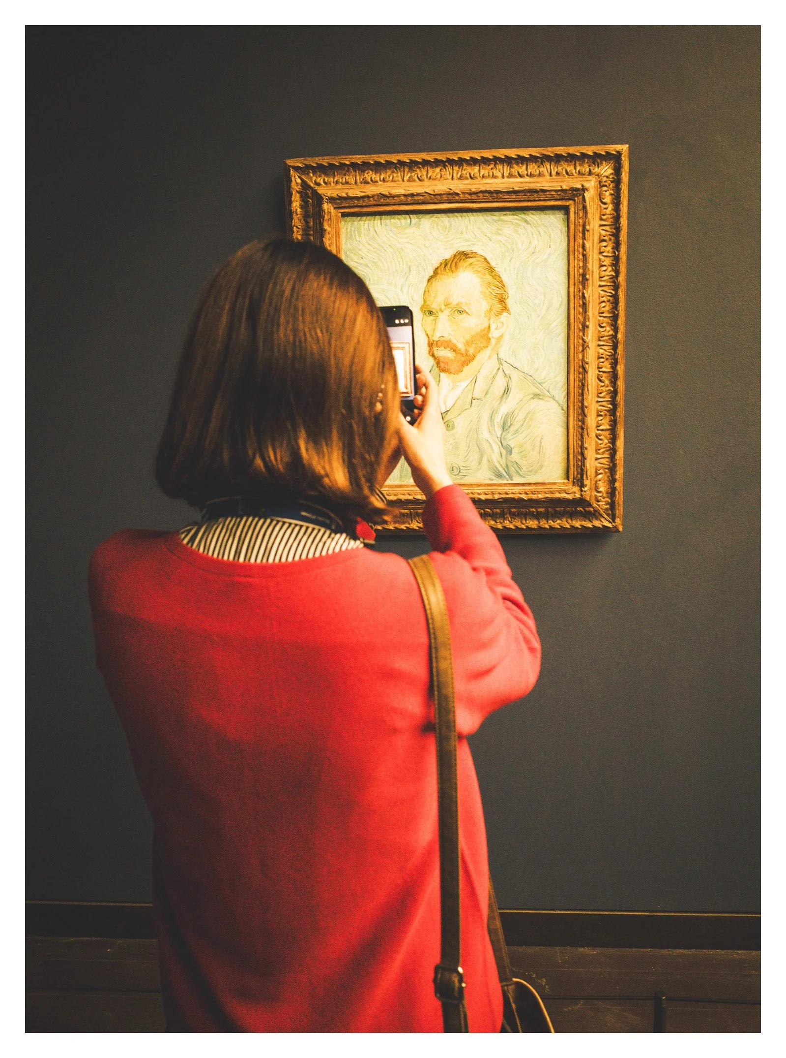 A woman taking a photo of a framed painting of Vincent van Gogh with her smartphone in an art gallery.