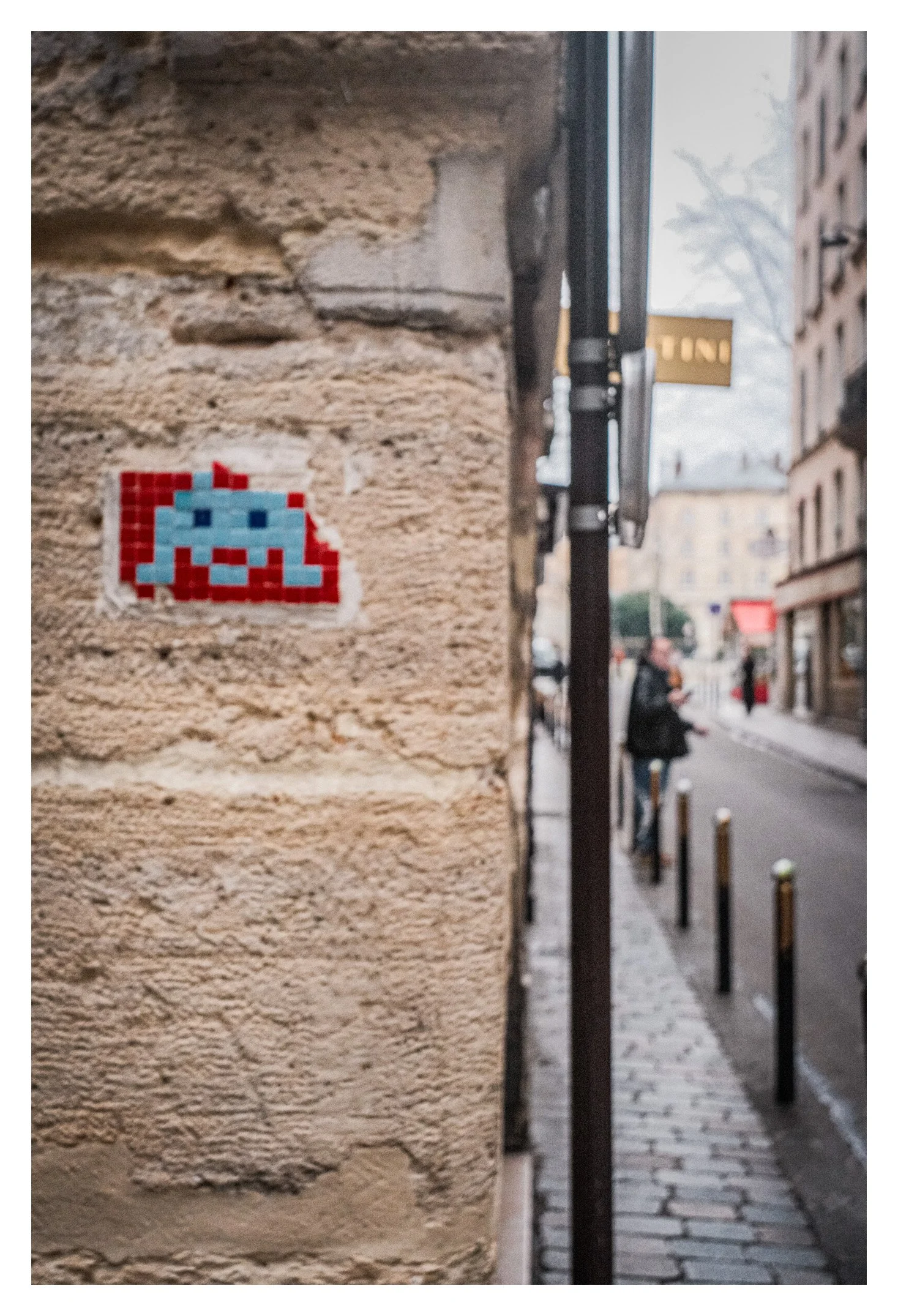 Pixel art depiction of a gray alien in red and blue colors on a brick wall in an urban street scene with pedestrians and buildings.