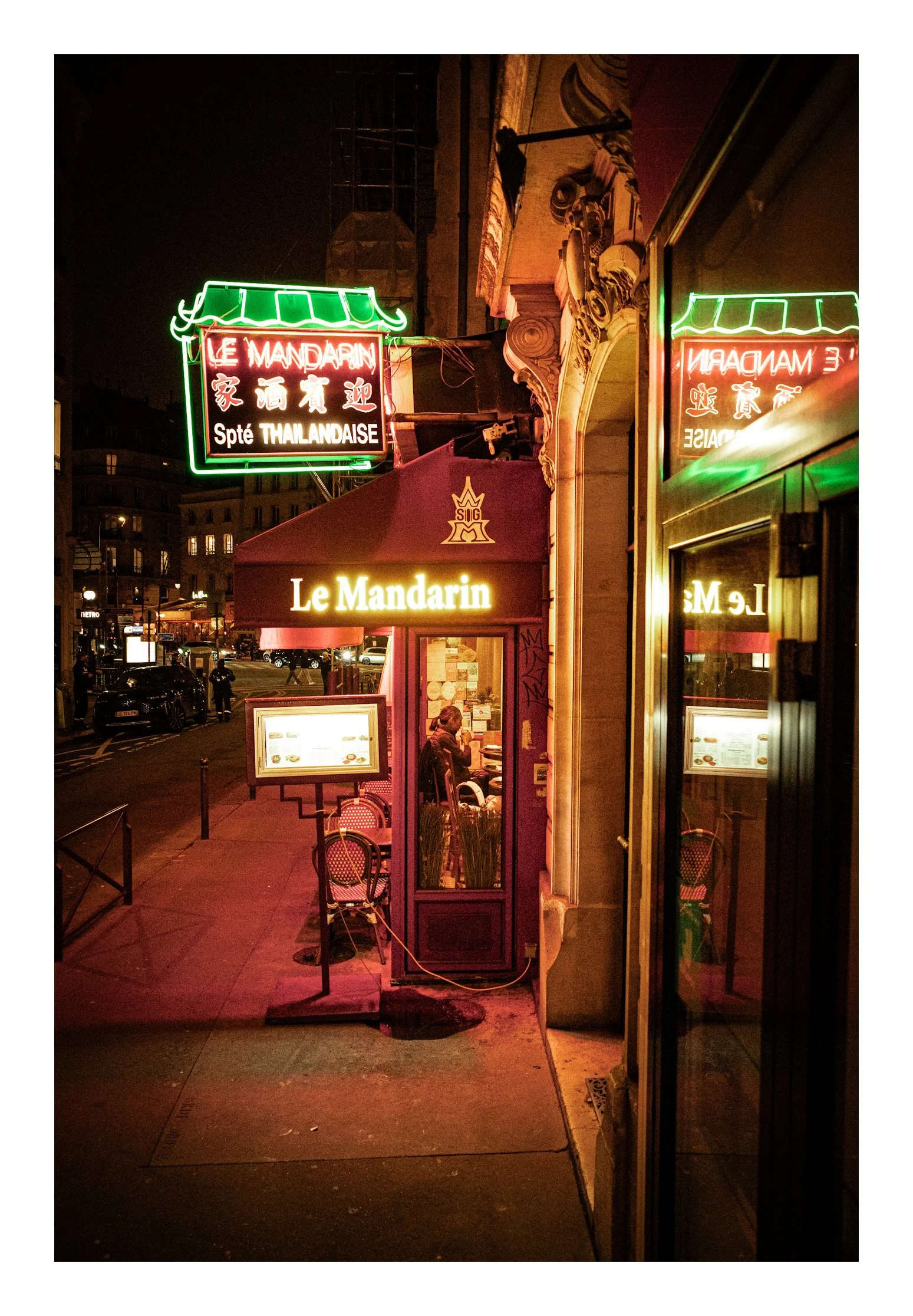 Night view of a Thai restaurant with neon signs reading 'Le Mandarin' and 'Spte Talandaise,' with outdoor seating and a person sitting inside.