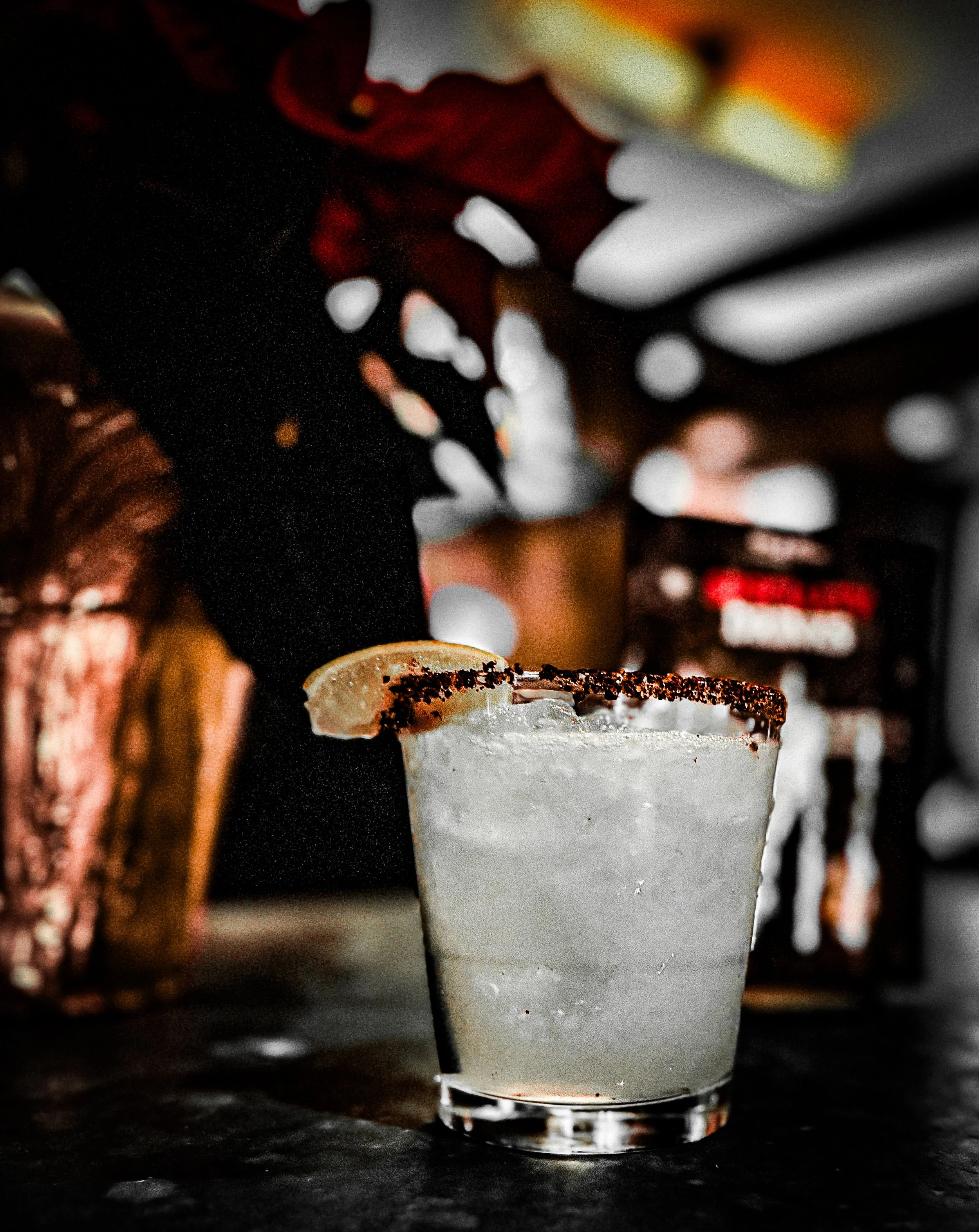 Close-up of a margarita cocktail glass with a salted rim and lemon wedge, on a dark bar counter, with blurry background of bottles and bar decor.