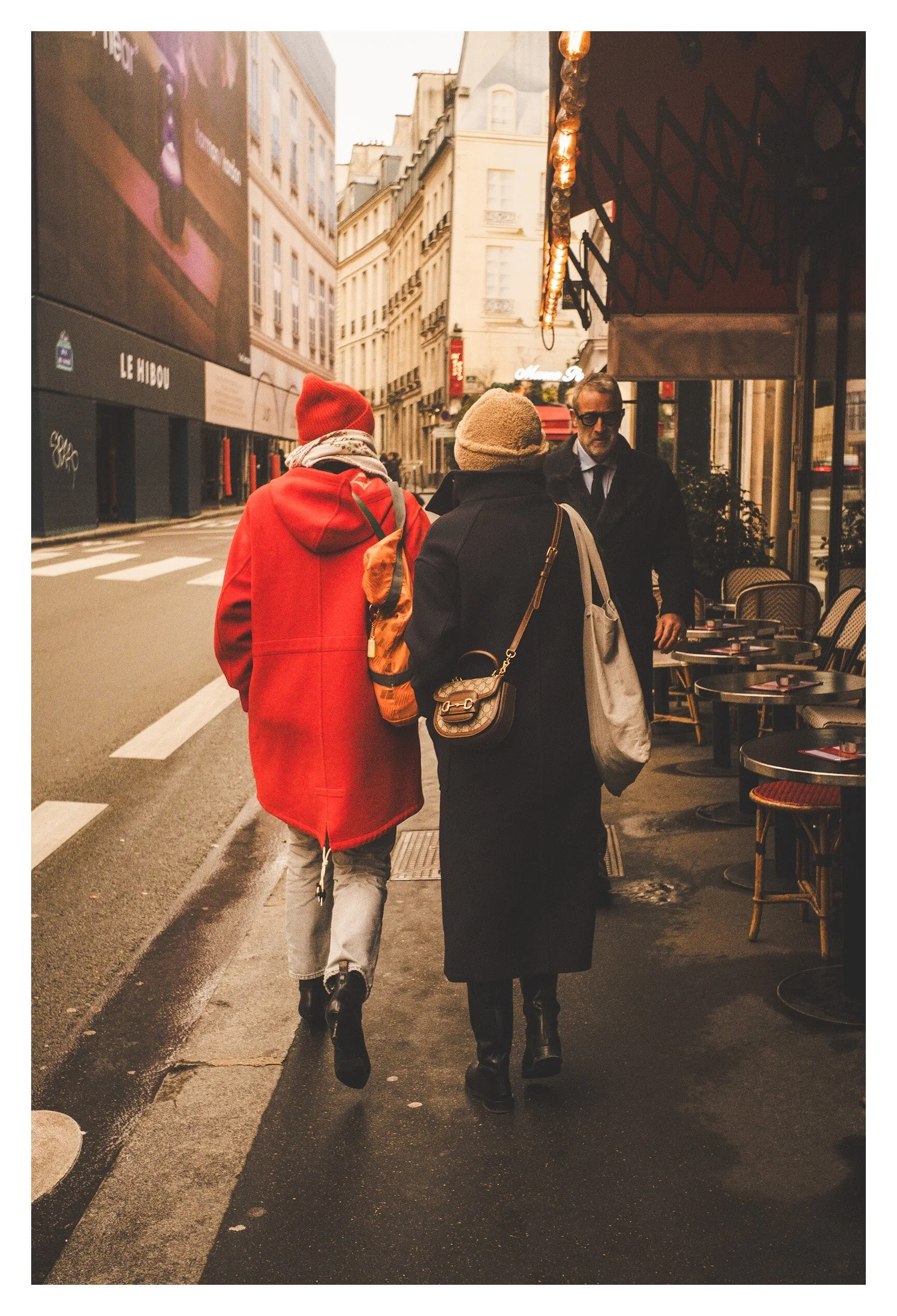 Three people walking down a city street on a cloudy day, with two of them seen from the back and one man facing forward. One person wears a red coat and beanie, the other in a dark coat with a beige hat, and the man in front wears sunglasses and a bl