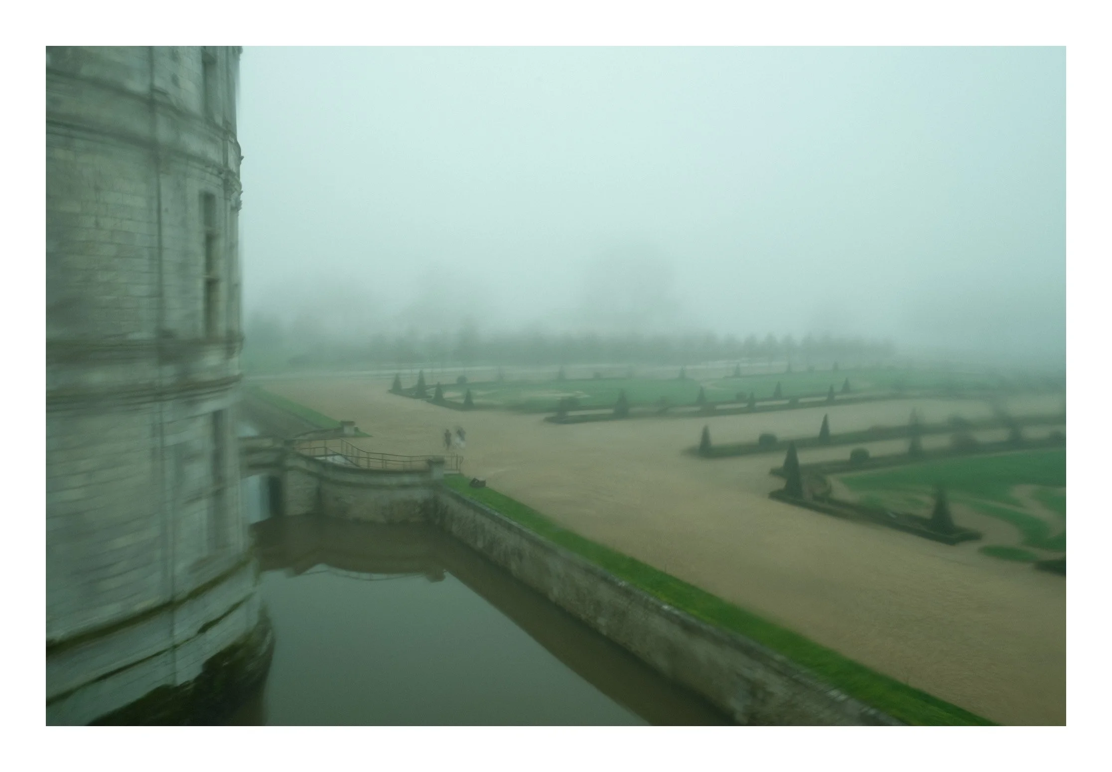Foggy view from a castle window showing a courtyard with pathways, trimmed hedges, and two small figures walking in the distance.