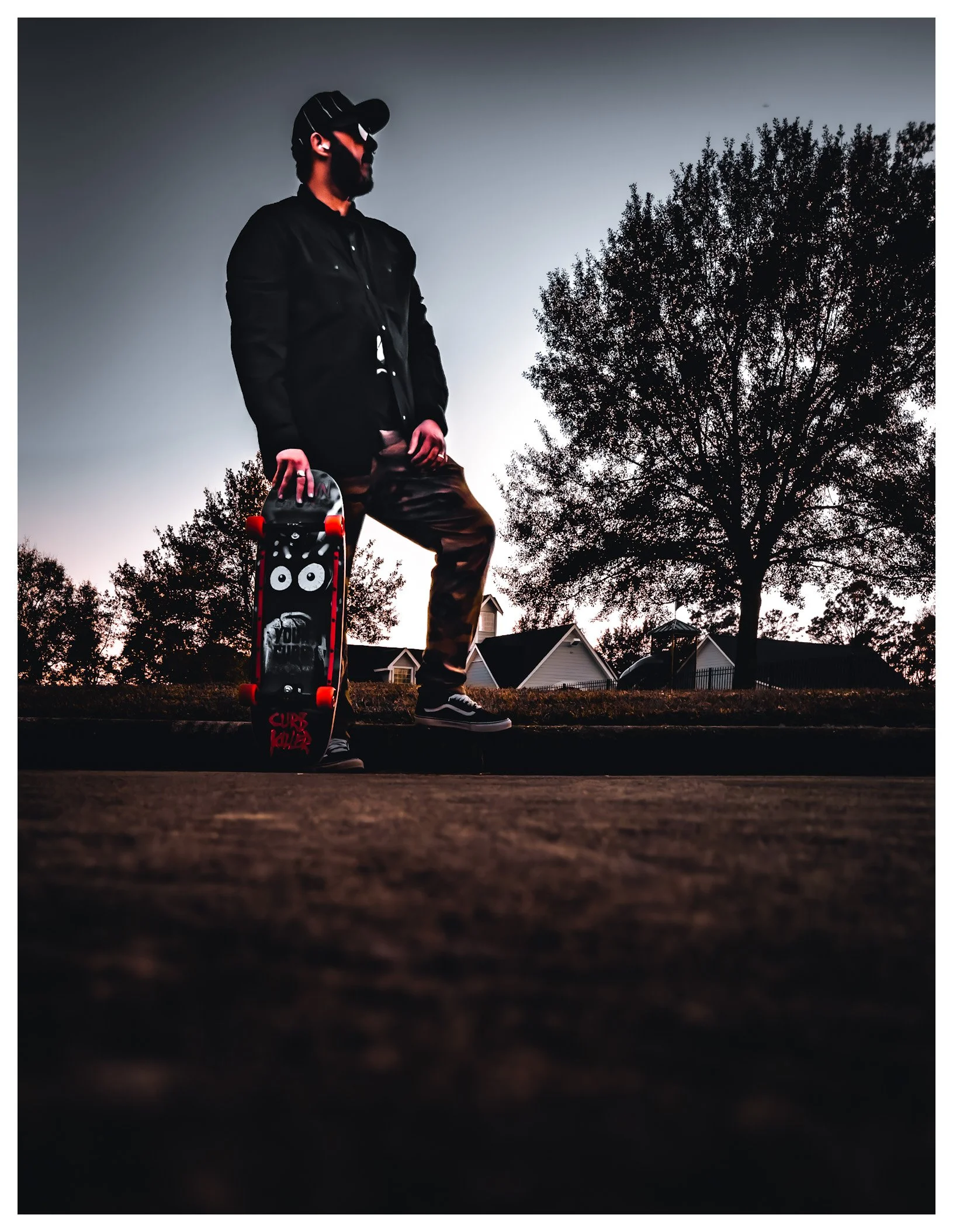 A man standing outdoors at dusk, holding a skateboard, wearing a black jacket, camouflage pants, black shoes, and a black cap, with trees and houses in the background.