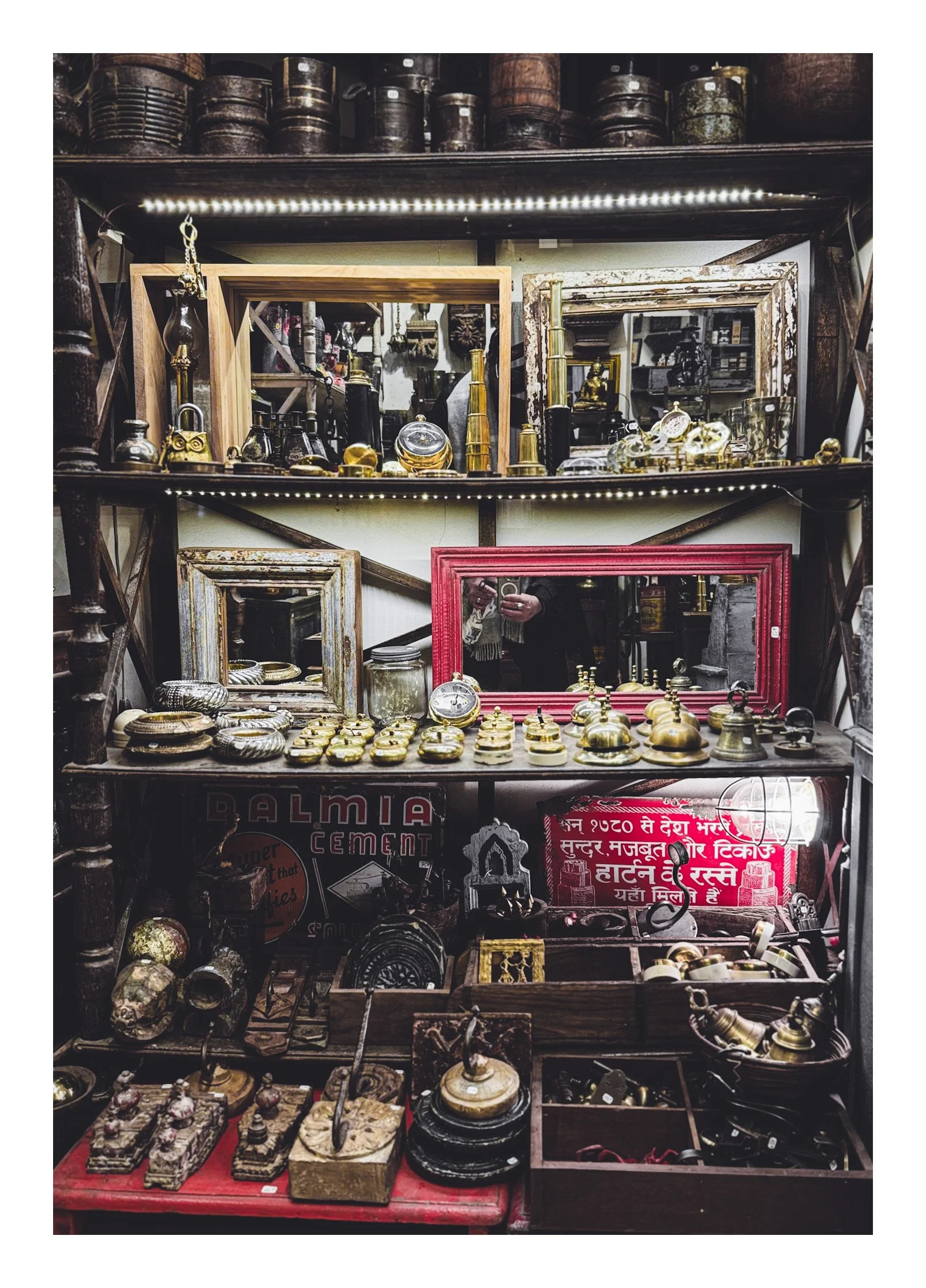 A cluttered antique shop display with vintage mirrors, brass and metal decorative items, small clocks, and various old trinkets on wooden shelves and tables.