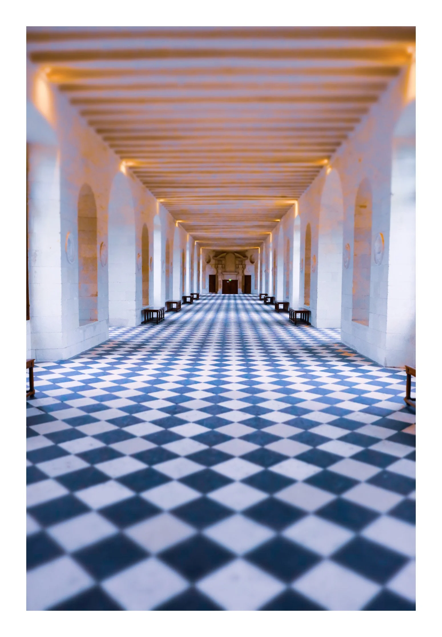 Long hallway with checkered black and white floor, stone walls with arches, and wooden ceiling beams, leading to a distant doorway.