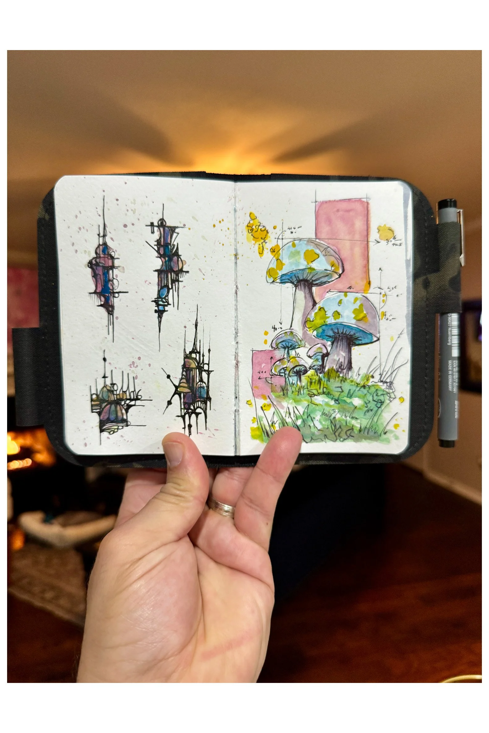 A sketchbook with abstract and whimsical drawings of fungi and mushrooms, with colorful ink and watercolor, held in a person's hand in front of a cozy room with a fireplace and warm lighting.