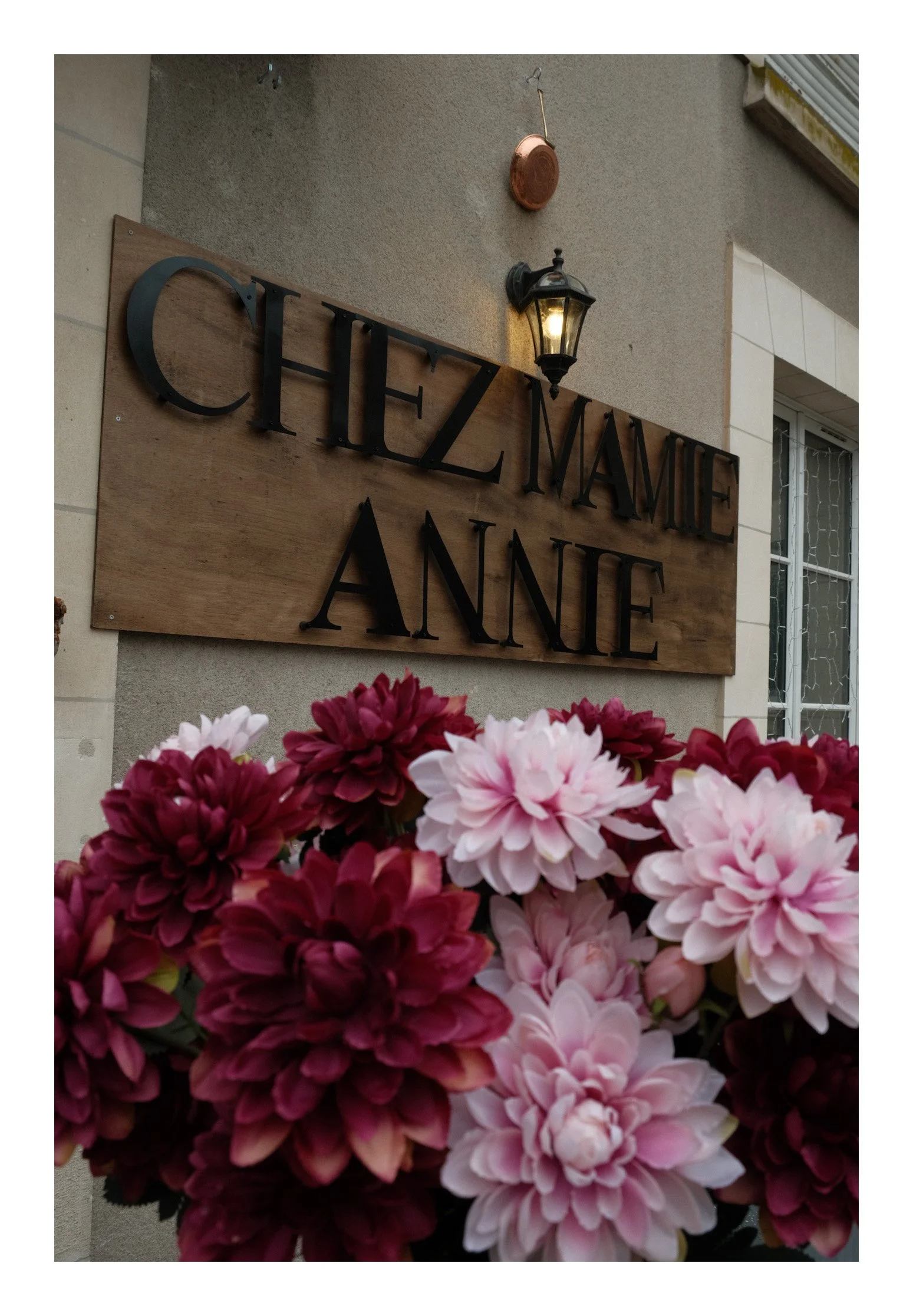 A wooden sign with black text reads 'Cinzannie Anne,' mounted on a beige wall next to a window with decorative grid pattern. A black outdoor lantern with a lit bulb is above the sign, and pink and dark red dahlias are in the foreground.