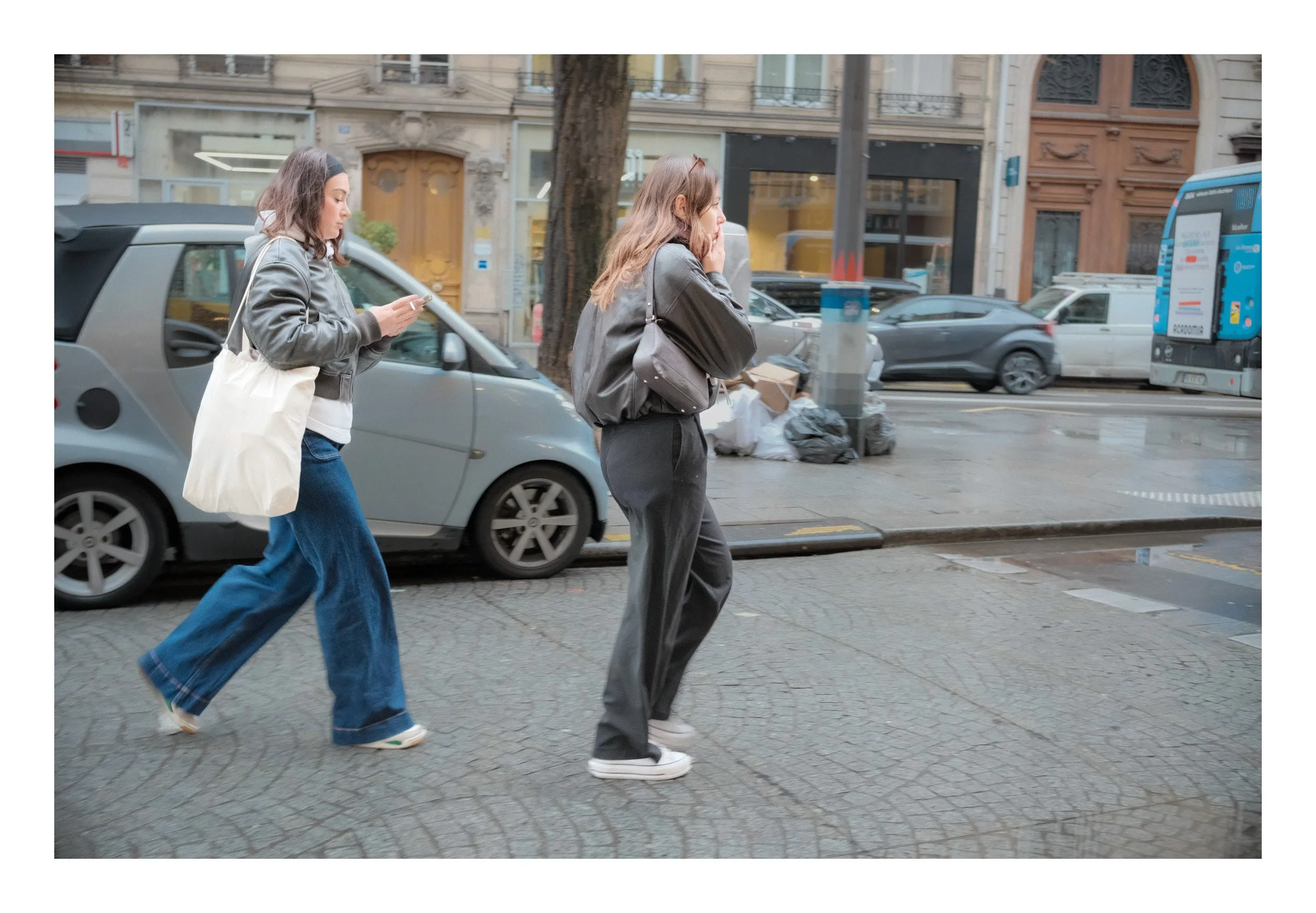 Two women walk on a city sidewalk, one is looking at her phone and the other is talking on a cellphone, with parked cars, a trash bag, and city buildings in the background.