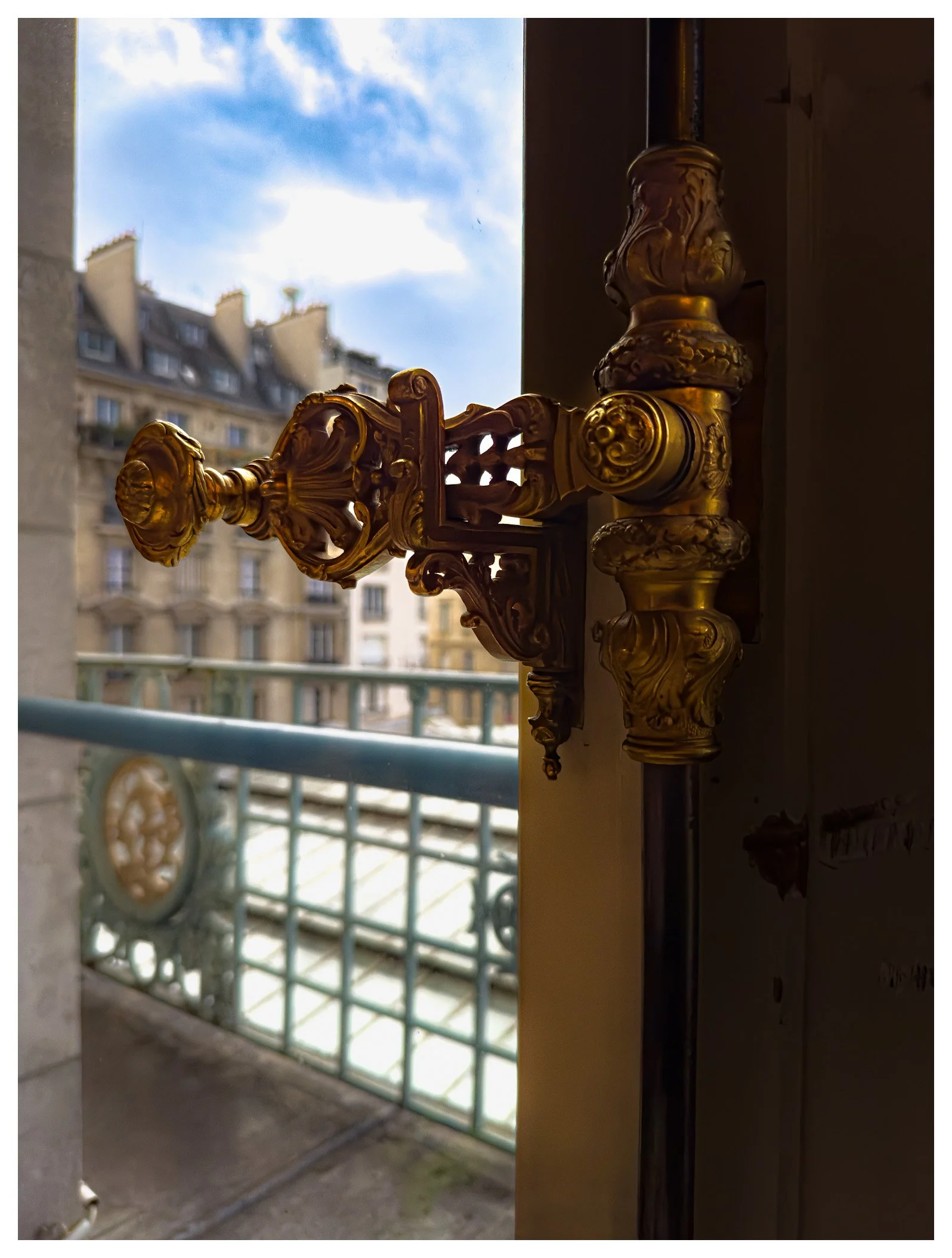 Close-up of an ornate, gold-colored decorative wall sconce on a window frame, with buildings and a cloudy sky visible outside.