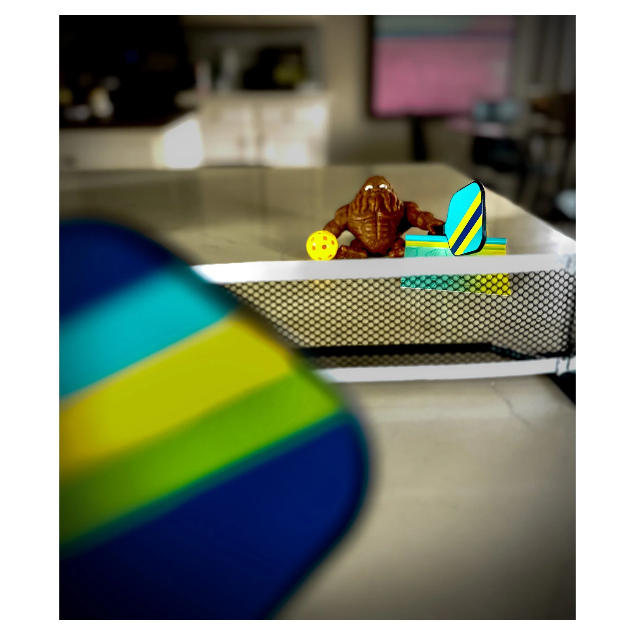 A collection of small toys on a desk, including a brown creature resembling a monster, a yellow die with red dots, and a small electronic device with a colorful striped design, with part of a blurred object in the foreground.