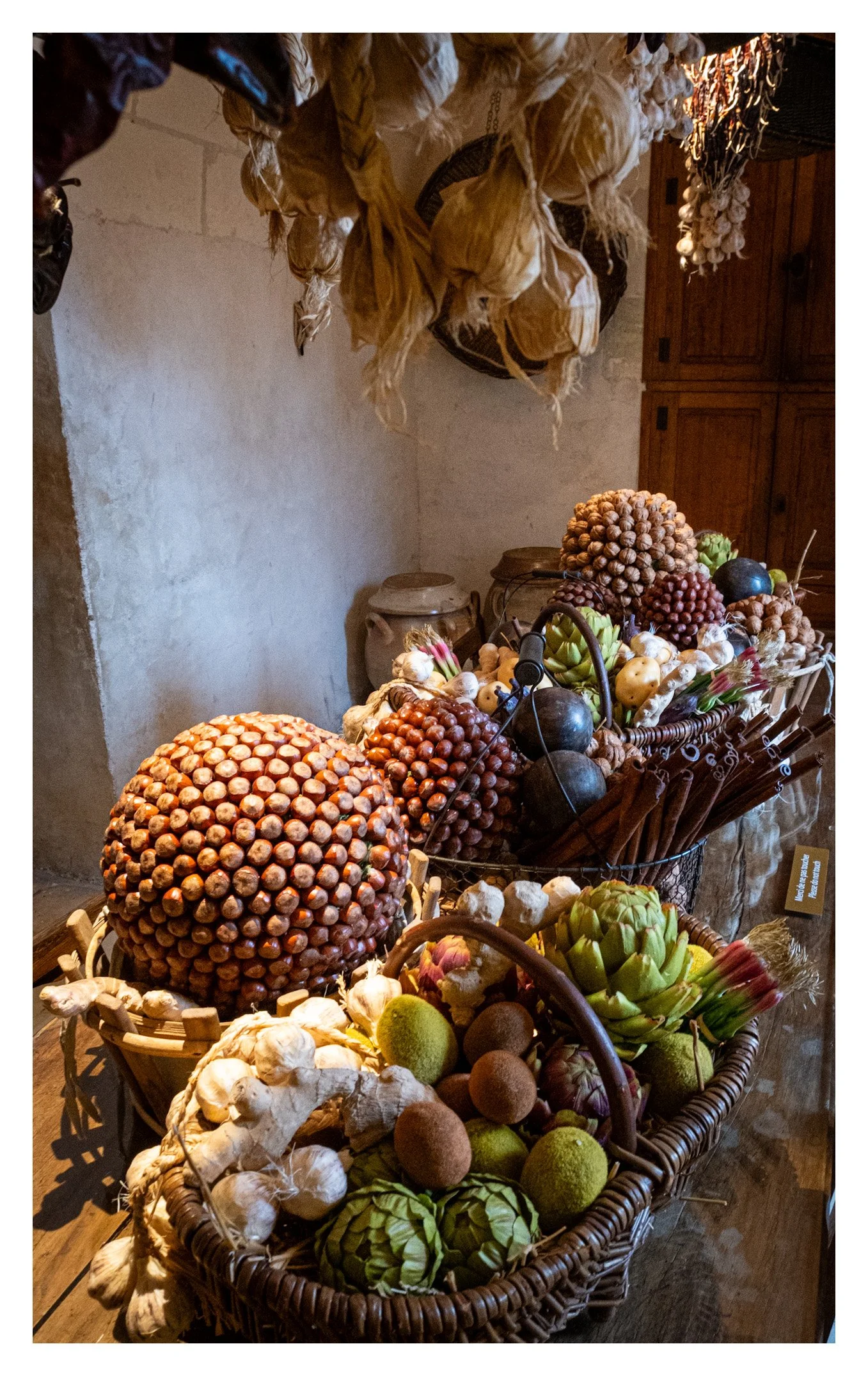 Basket of various vegetables and fruits, including artichokes, garlic, hazelnuts, and onions, on a wooden surface in a rustic kitchen setting.