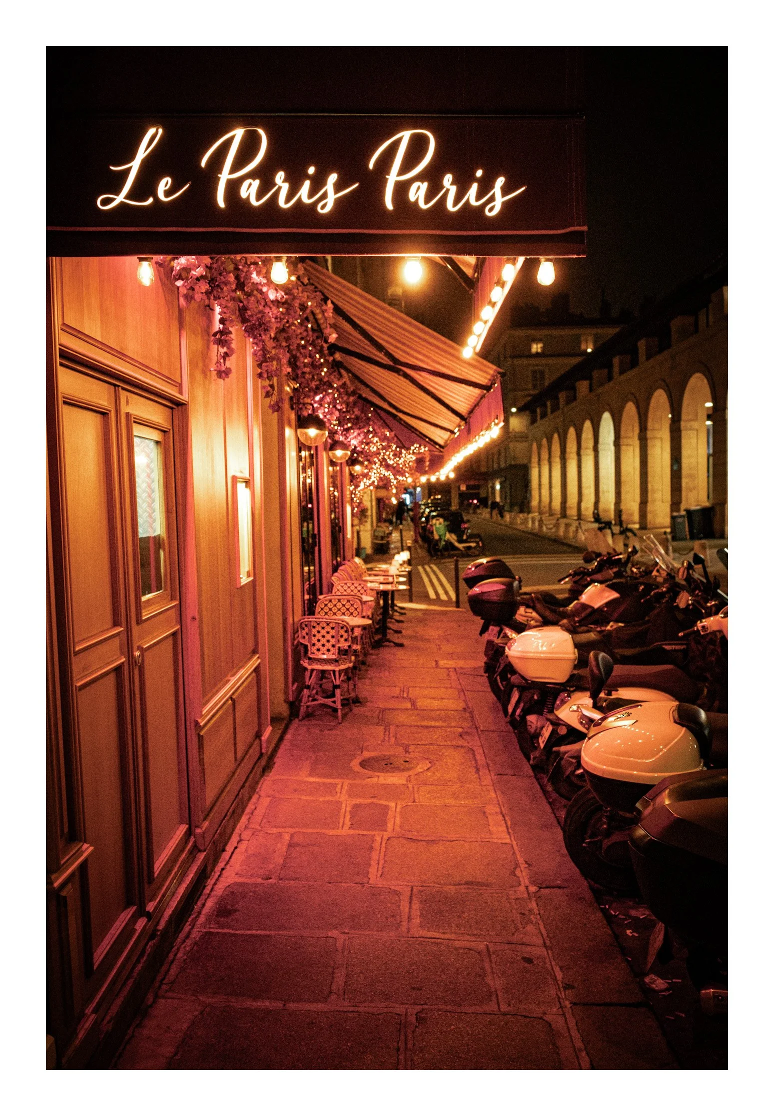 Night scene of a European-style cafe with outdoor seating, illuminated with warm string lights, pink flowers, and a sign reading 'Le Paris Paris'.