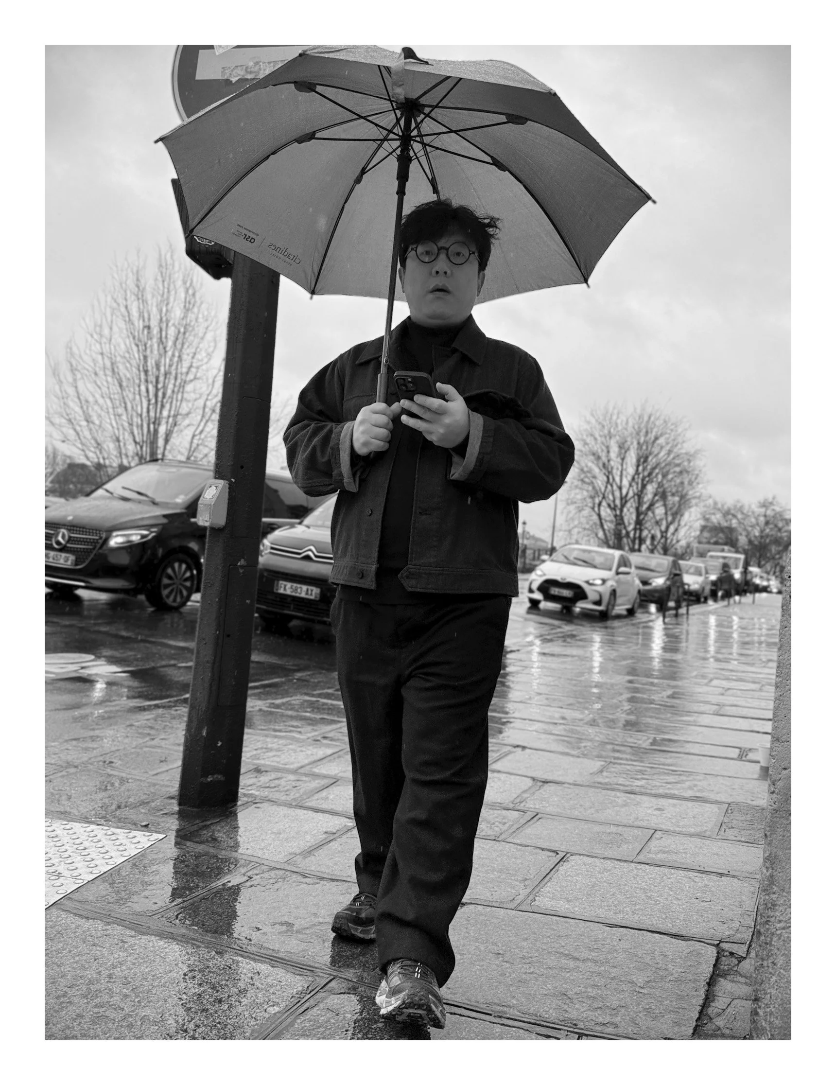 A man with glasses holding an umbrella and looking at his phone while walking on a wet street in rainy weather, with parked cars and leafless trees in the background.