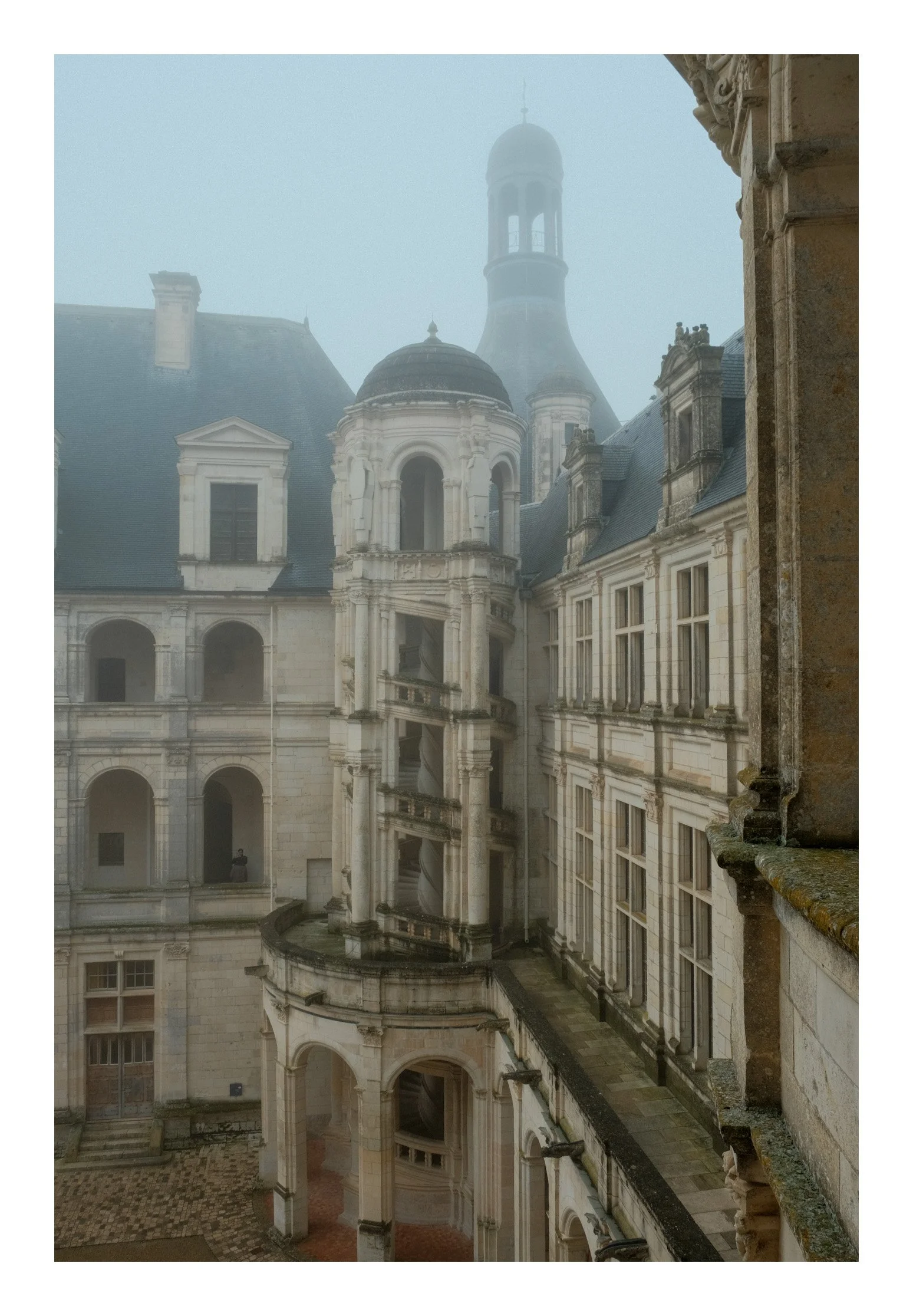 A historic castle or palace with stone walls and turrets, partially covered by fog, including a staircase on the exterior and arched windows.
