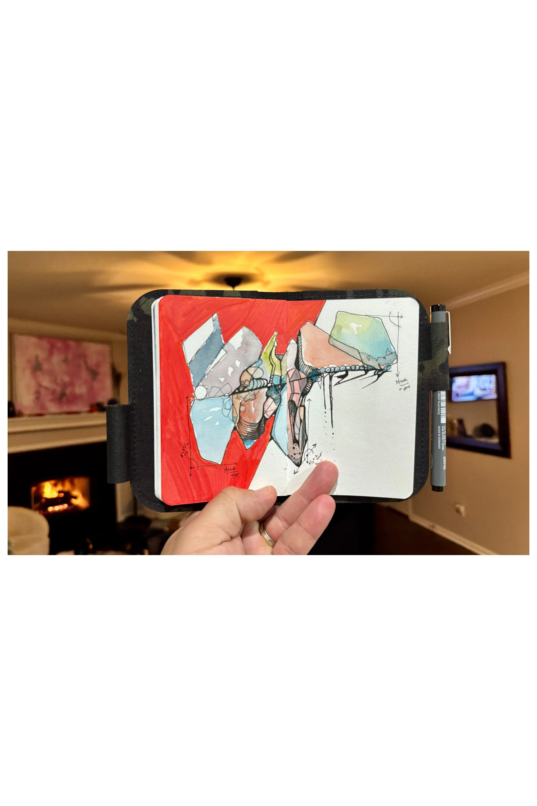 A hand holding a sketchbook open with a colorful abstract drawing inside. The background shows a cozy living room with a fireplace and a sliding door TV.