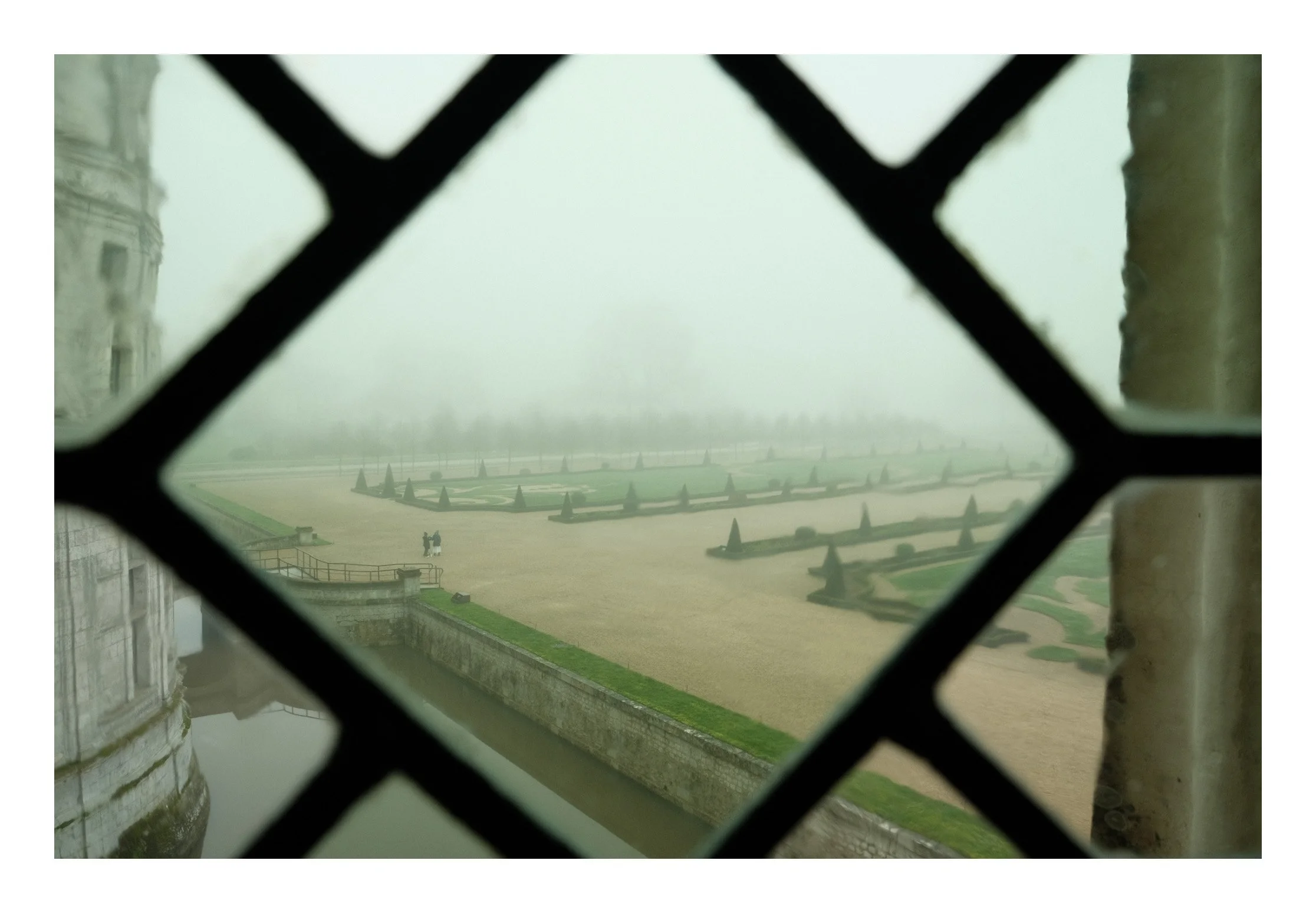 View of a foggy landscape with neatly landscaped gardens, spires, and two people walking, seen through a decorative iron grate.