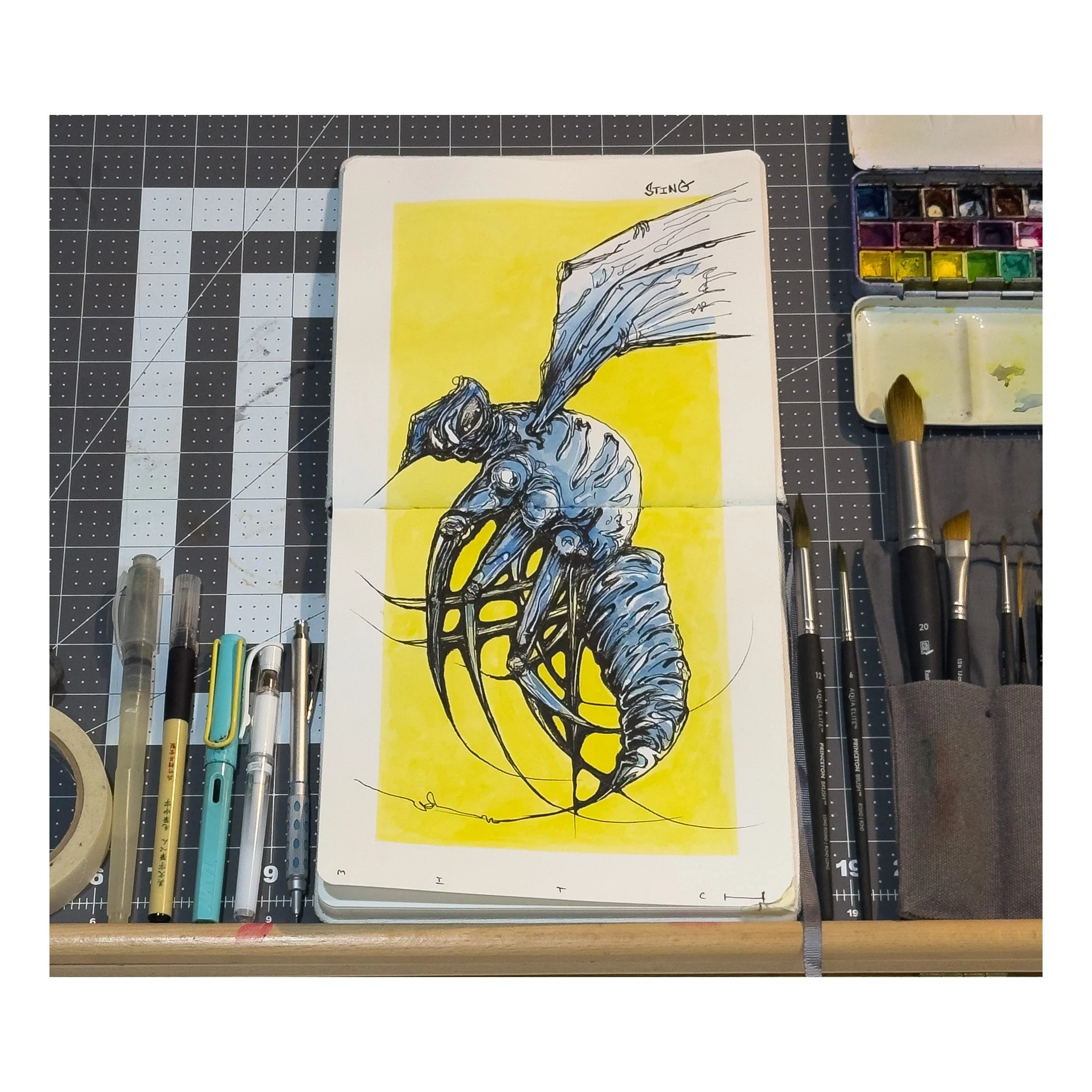 A drawing of a creature with a large, segmented, worm-like body and a humanoid upper half wearing goggles, set against a yellow background, with various art supplies on a cutting mat nearby.