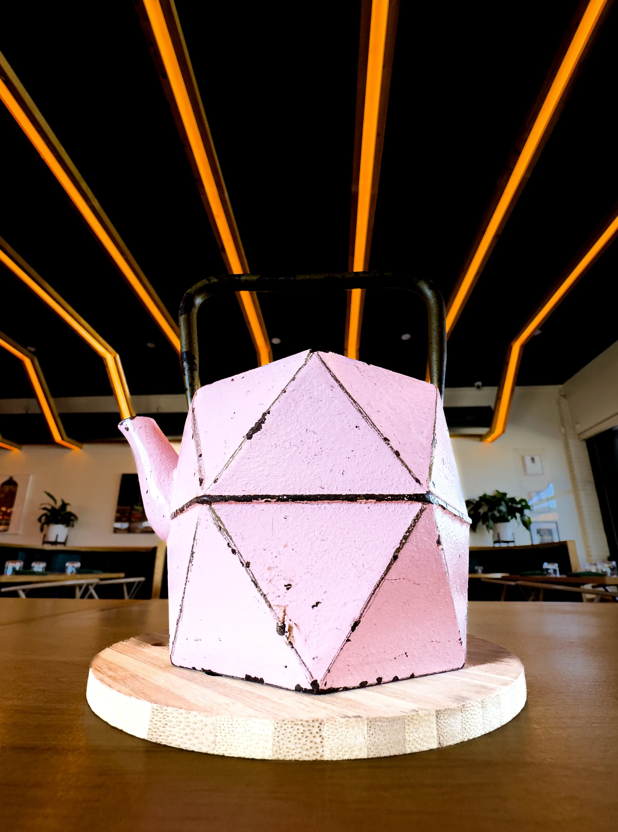 Pink teapot with black lines forming geometric shapes, placed on a round wooden base, inside a modern room with black ceiling, orange neon lights, and plants in the background.