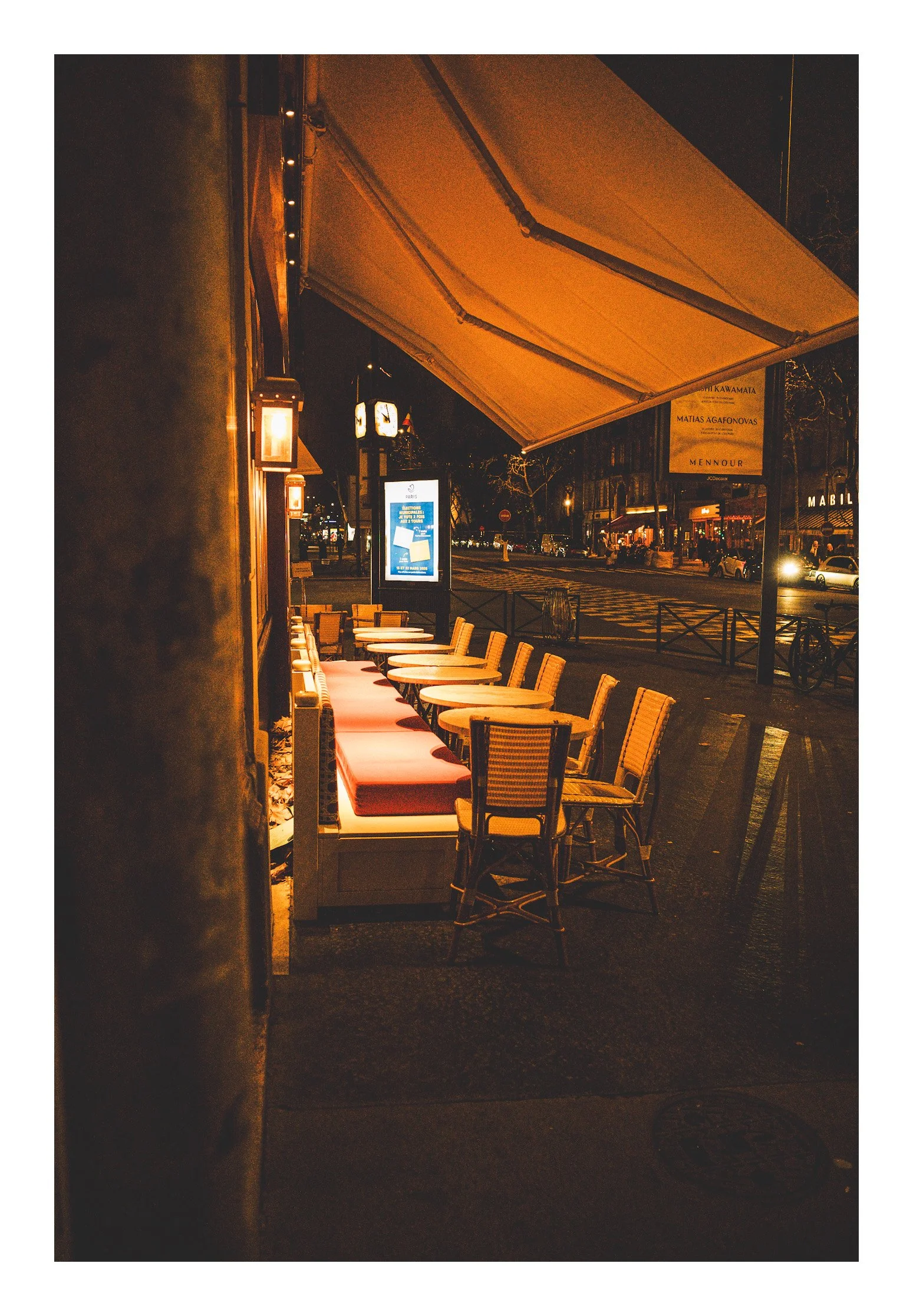 Empty outdoor café seating area at night, with chairs and tables along a city sidewalk illuminated by warm lighting.