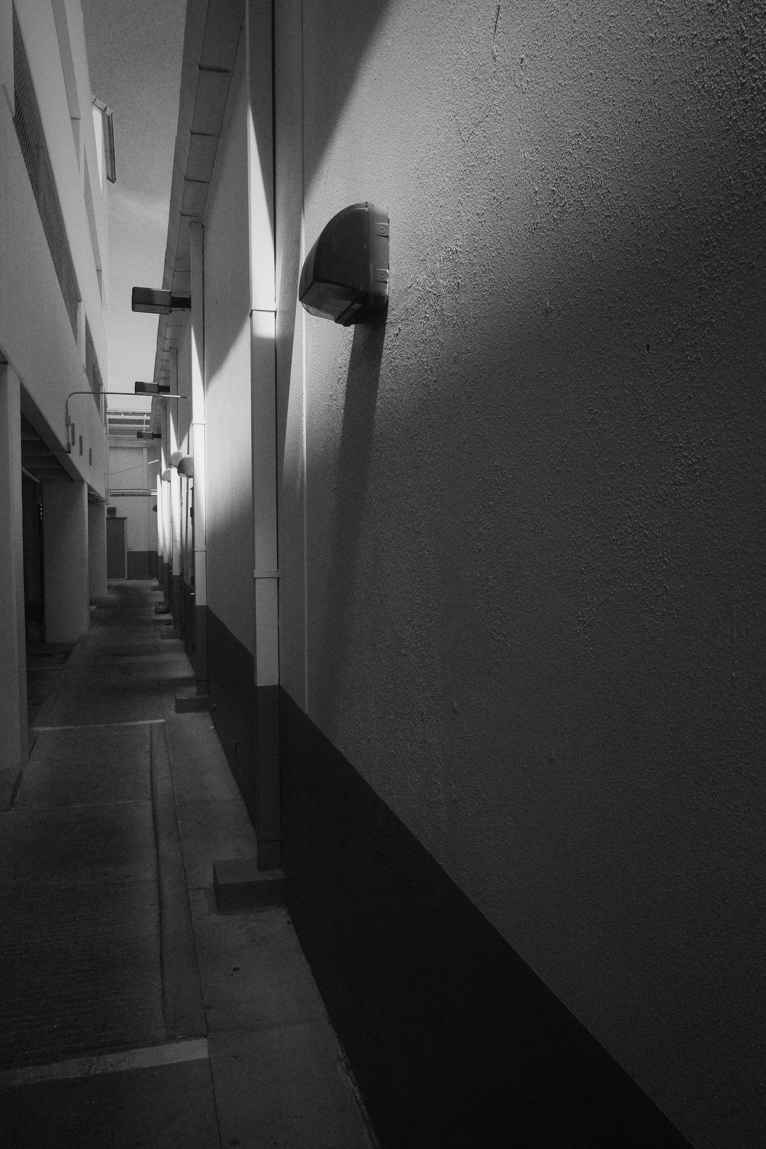 Black and white photo of an alleyway with angled shadows cast on the wall, showing air conditioning units and other building features.