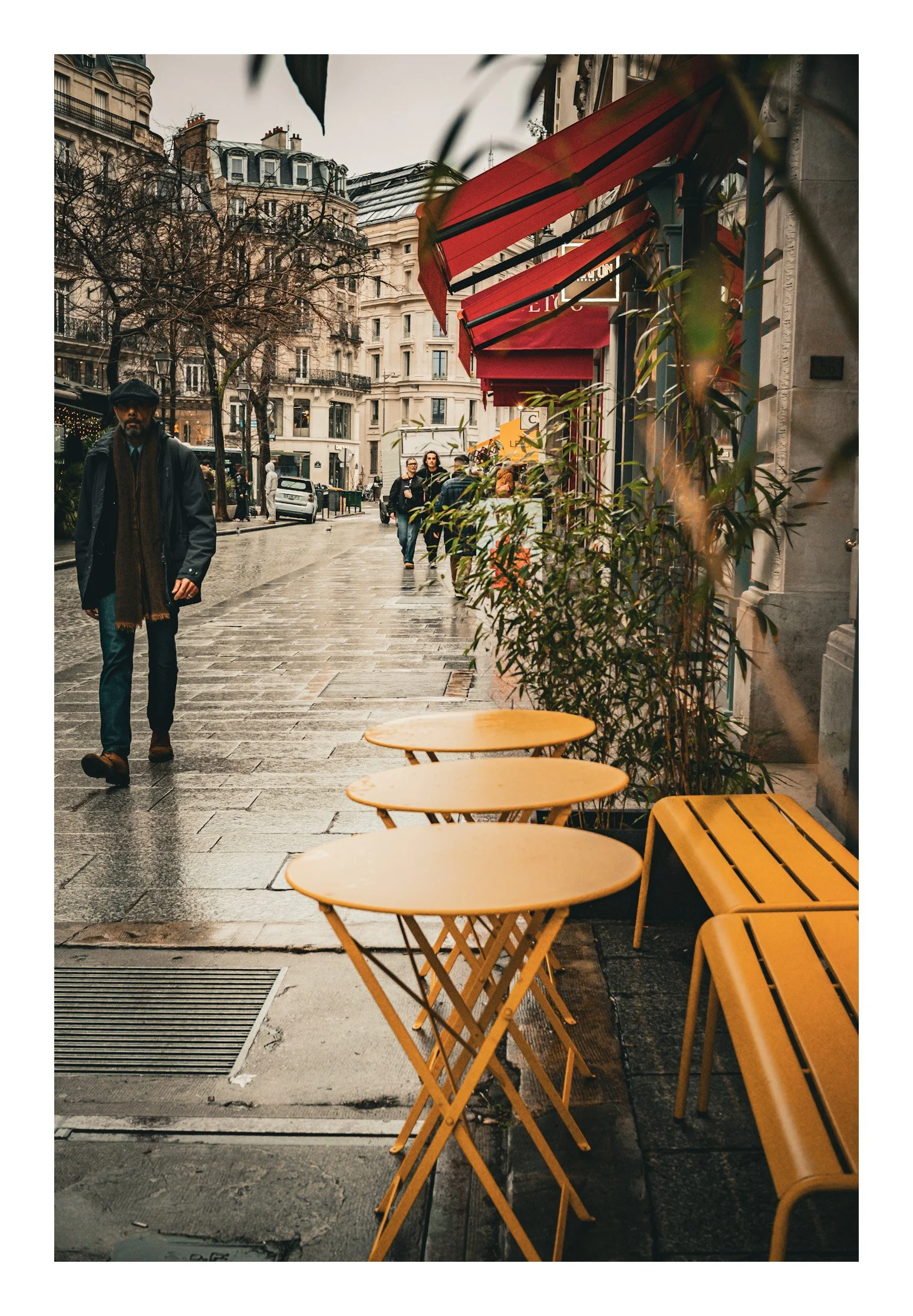 Empty outdoor café tables and benches on a sidewalk in an urban setting with pedestrians walking on a rainy day, Parisian buildings in the background.