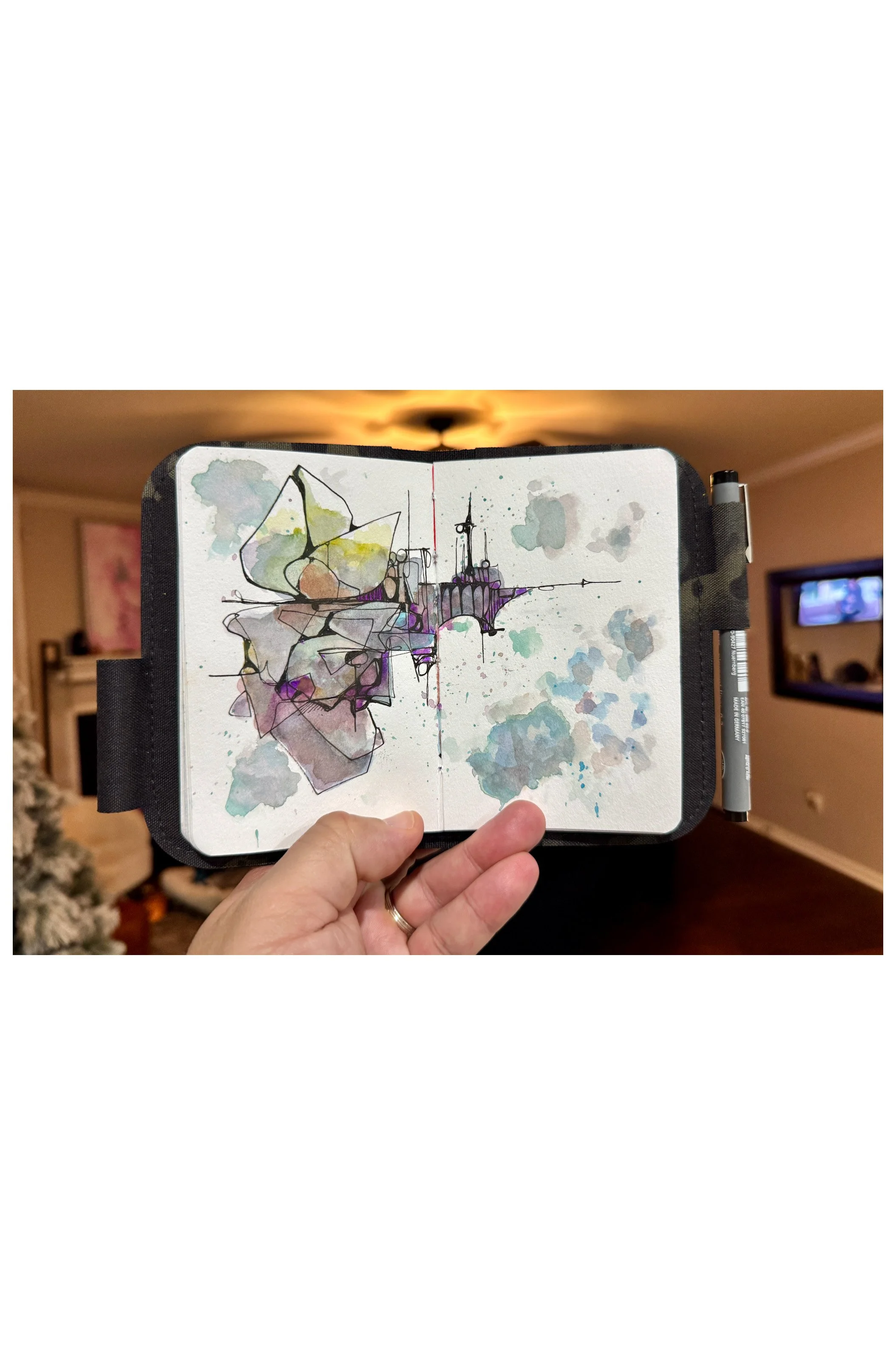 Open sketchbook with abstract watercolor drawing of a floating city and clouds, held in a person's hand, in a living room with Christmas tree and television.