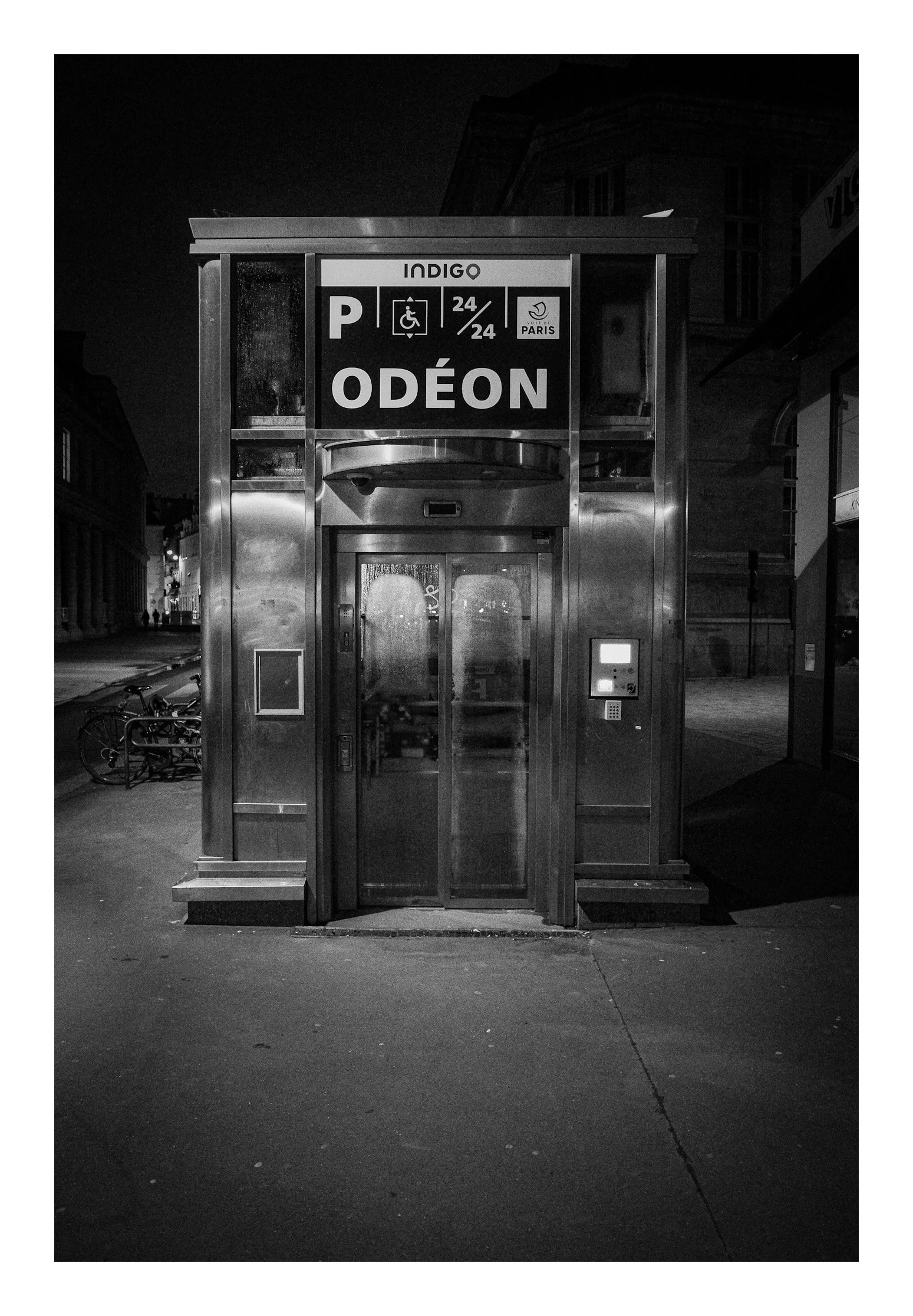 A black and white photo of a small public telephone booth in an urban setting at night.