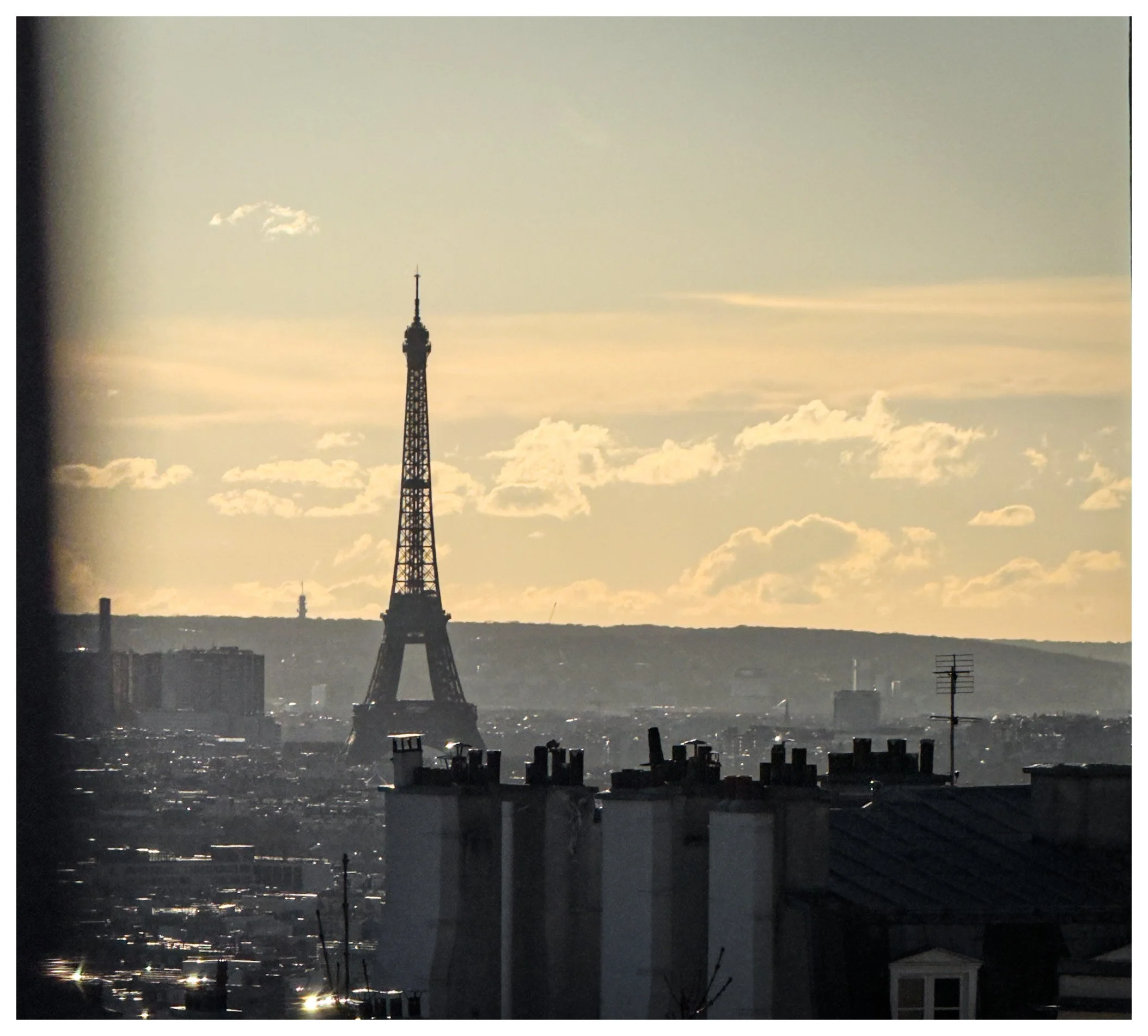 A sunset view of Paris with the Eiffel Tower in the distance, seen from a balcony with the rooftops of surrounding buildings in the foreground.