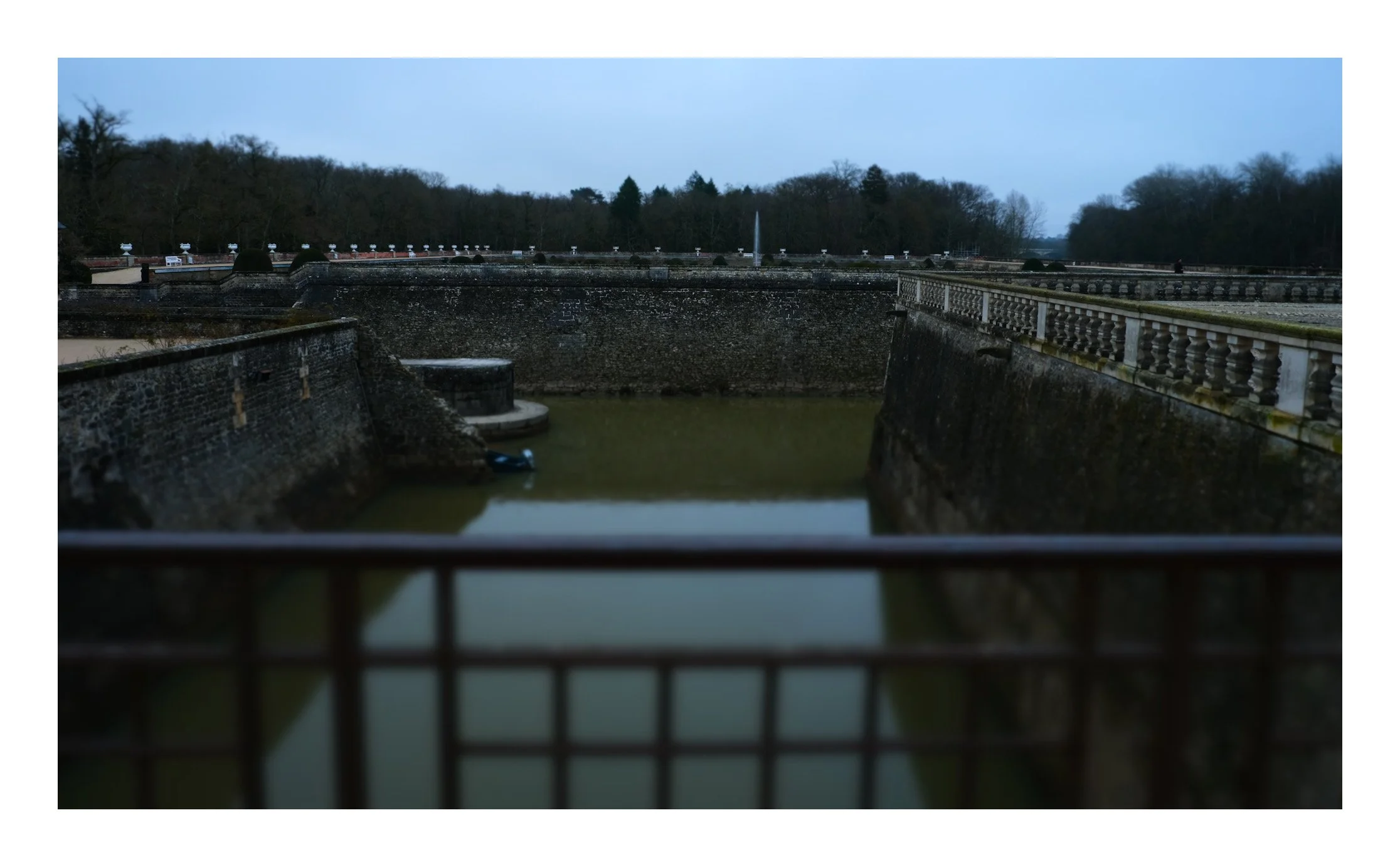 View of a historic stone lock with water inside and surrounding stone walls, trees in the background, and cloudy sky.