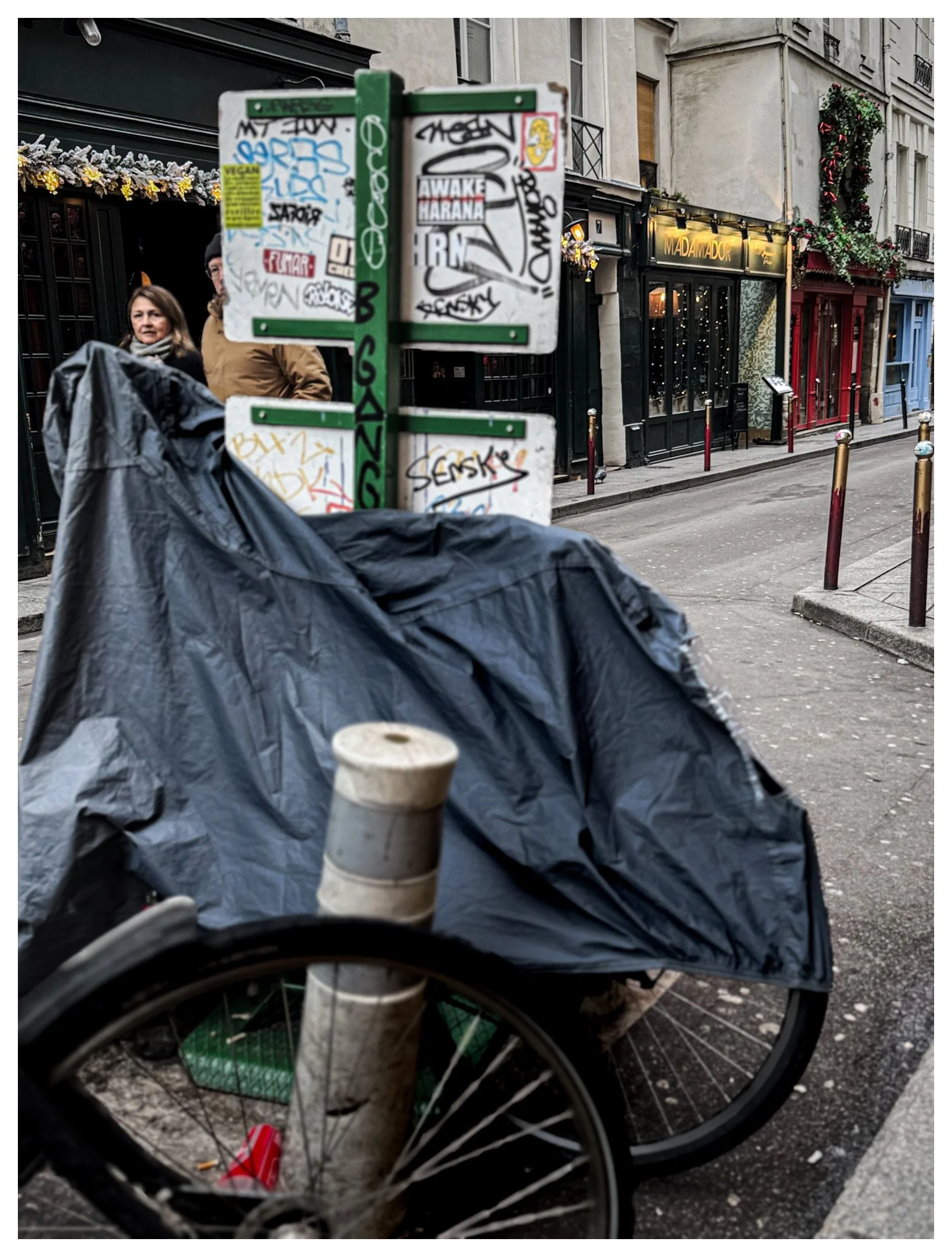 A street scene with a bicycle covered by a black plastic bag in the foreground, graffiti-covered signs on a pole, and a woman standing near a building decorated with holiday wreaths and flowers in the background.