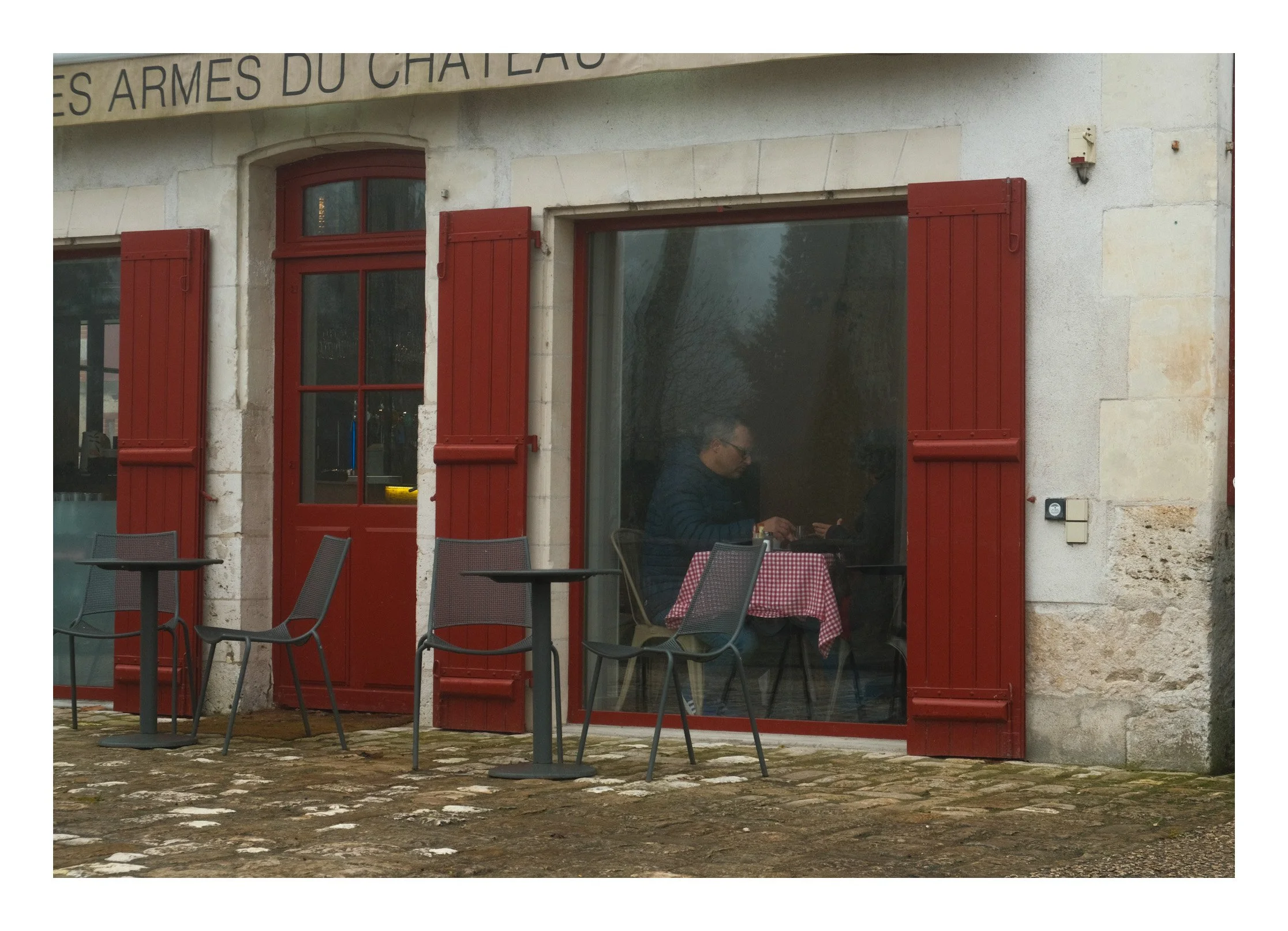 A man sitting inside a restaurant at a table with a red checkered tablecloth, seen through large glass windows with red wooden shutters outside. The exterior has a stone wall and cobblestone ground, with outdoor chairs and tables.