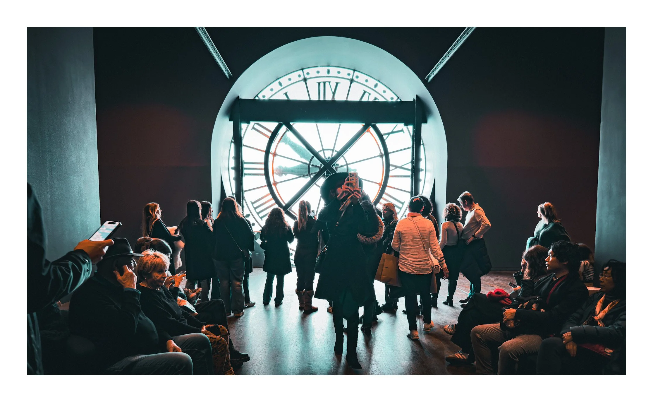 People observing a large clock with Roman numerals at an indoor location, some sitting and others standing, with one person taking a photo.
