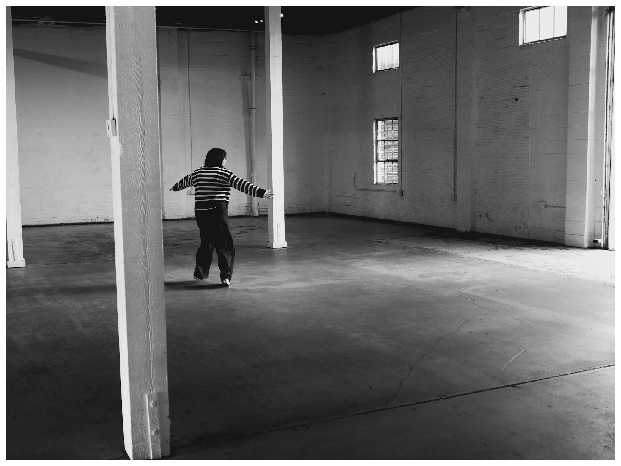 A person walking alone inside an empty industrial-style room with white brick walls, concrete floor, and high ceilings, lit by natural light from two windows.