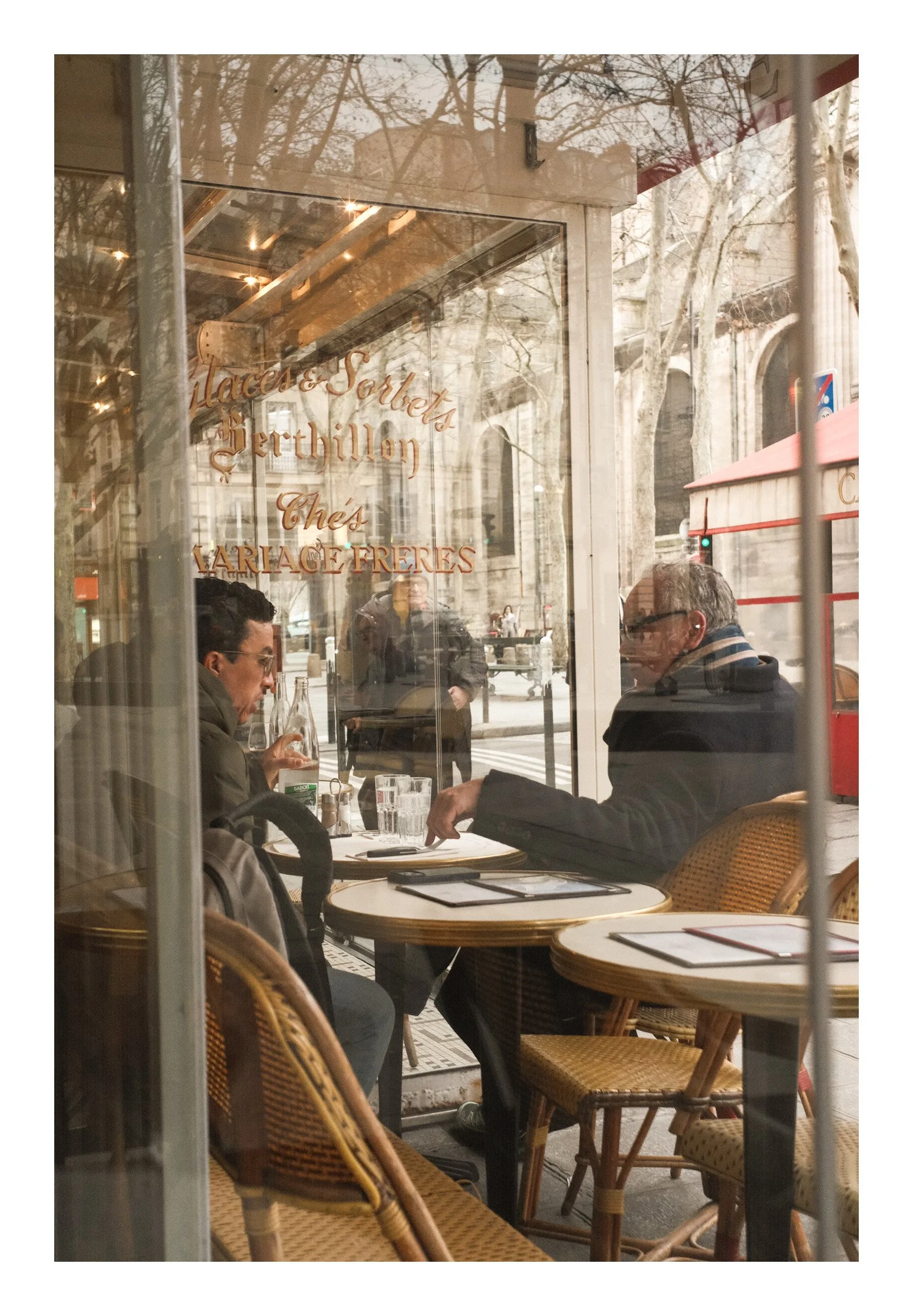 View through a restaurant window showing two men seated at a table with menus, glassware, and a bottle, with a street scene and pedestrians outside.