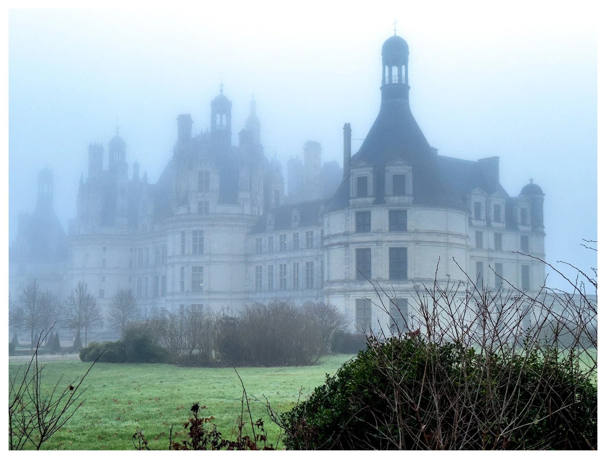 A foggy view of a historic castle with multiple towers and turrets, partially obscured by mist, with a grassy lawn and bushes in the foreground.