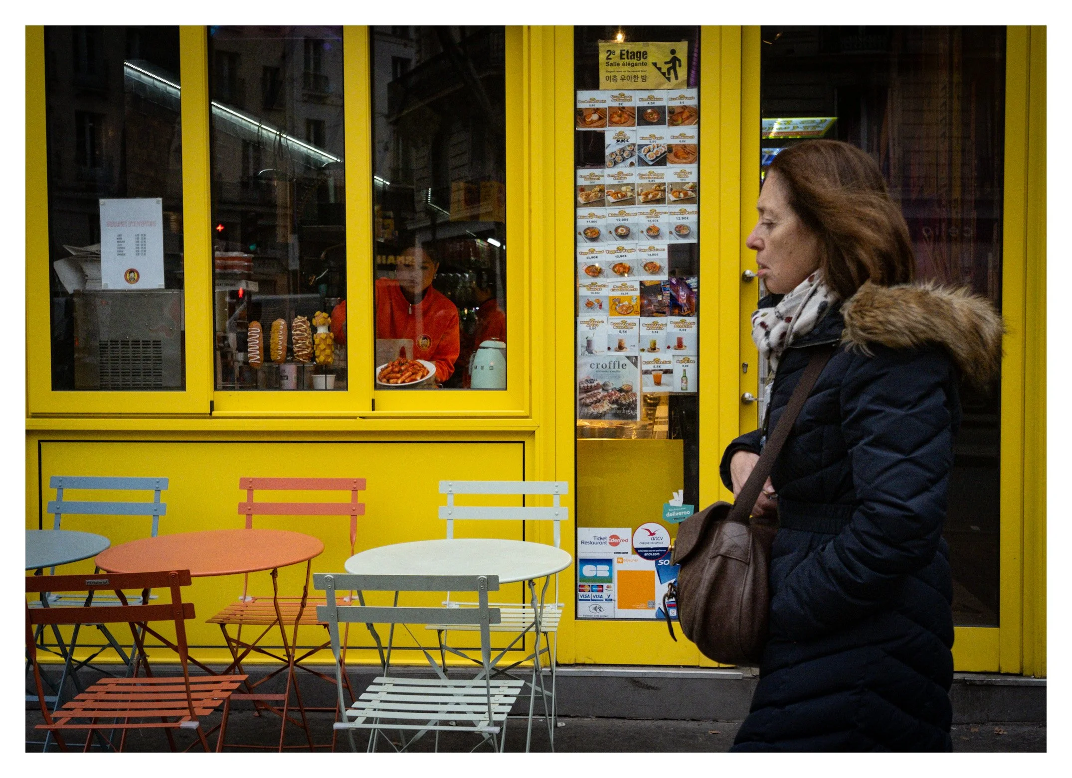 A woman with brown hair in a black coat and scarf stands outside a bright yellow cafe, looking inside where a worker in a red jacket is preparing food. The cafe has outdoor seating with colorful metal chairs and tables, and a menu with various dishes