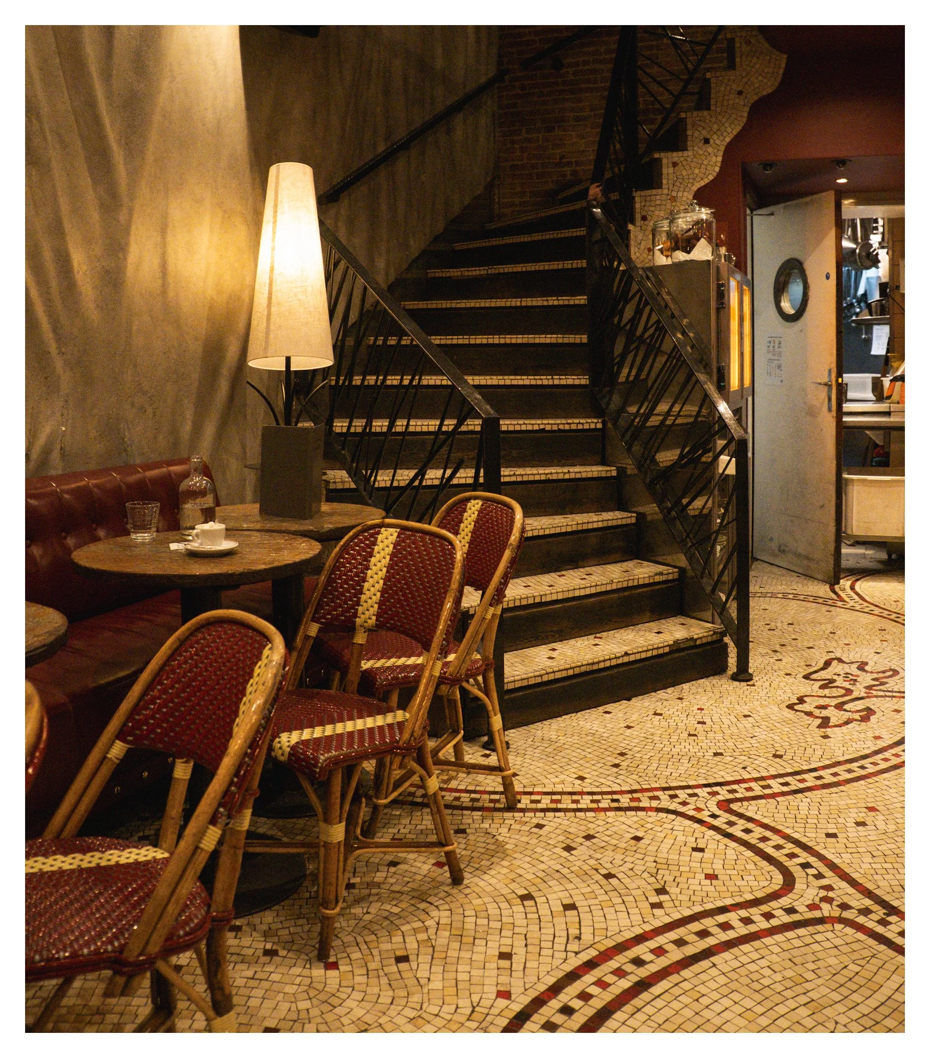 Inside a cozy restaurant with a mosaic tiled floor, a staircase with black railing, a table with glasses and coffee cups, and a lit floor lamp.