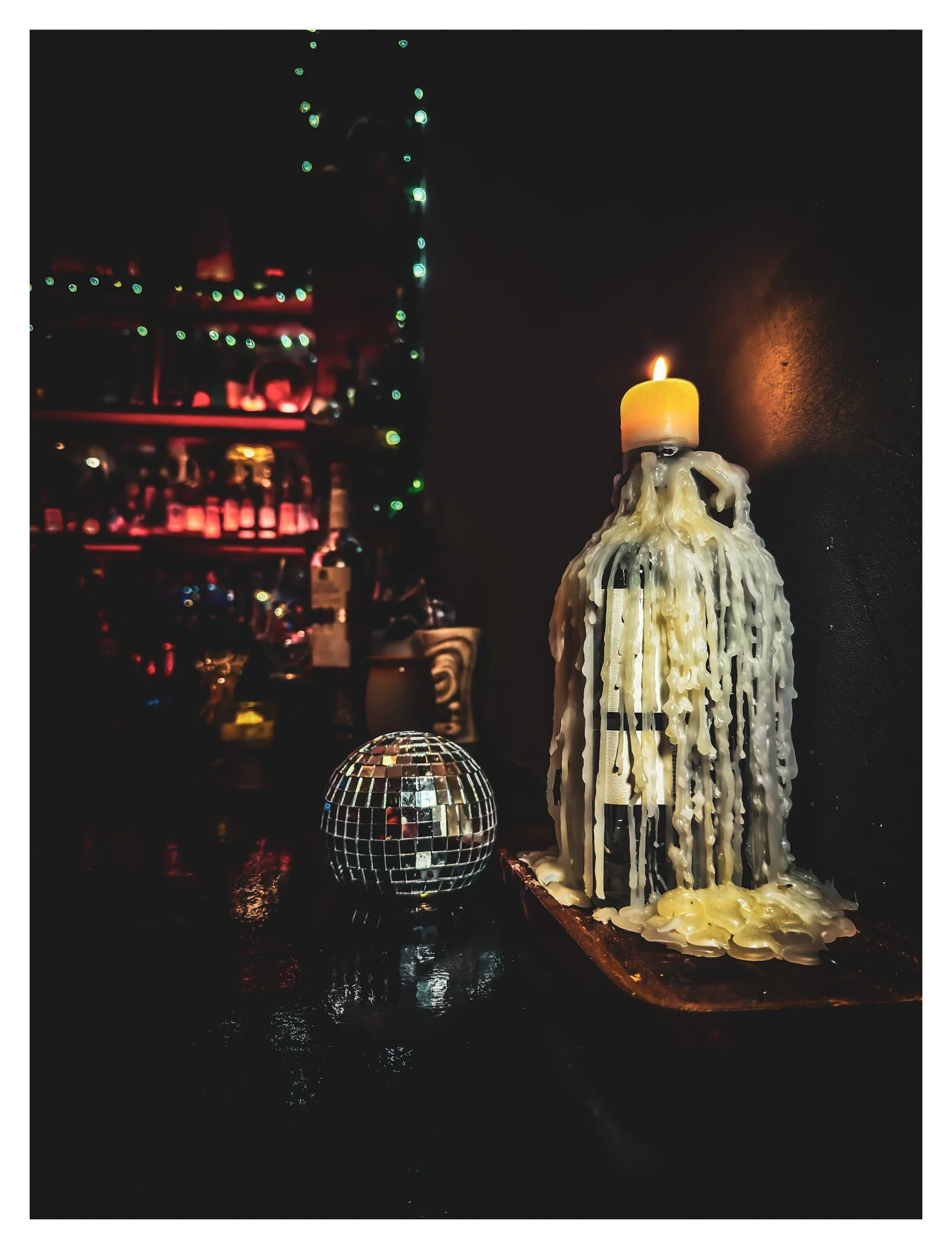 A lit candle melting over a tall candle holder, a small disco ball, and blurry colorful lights in the background.