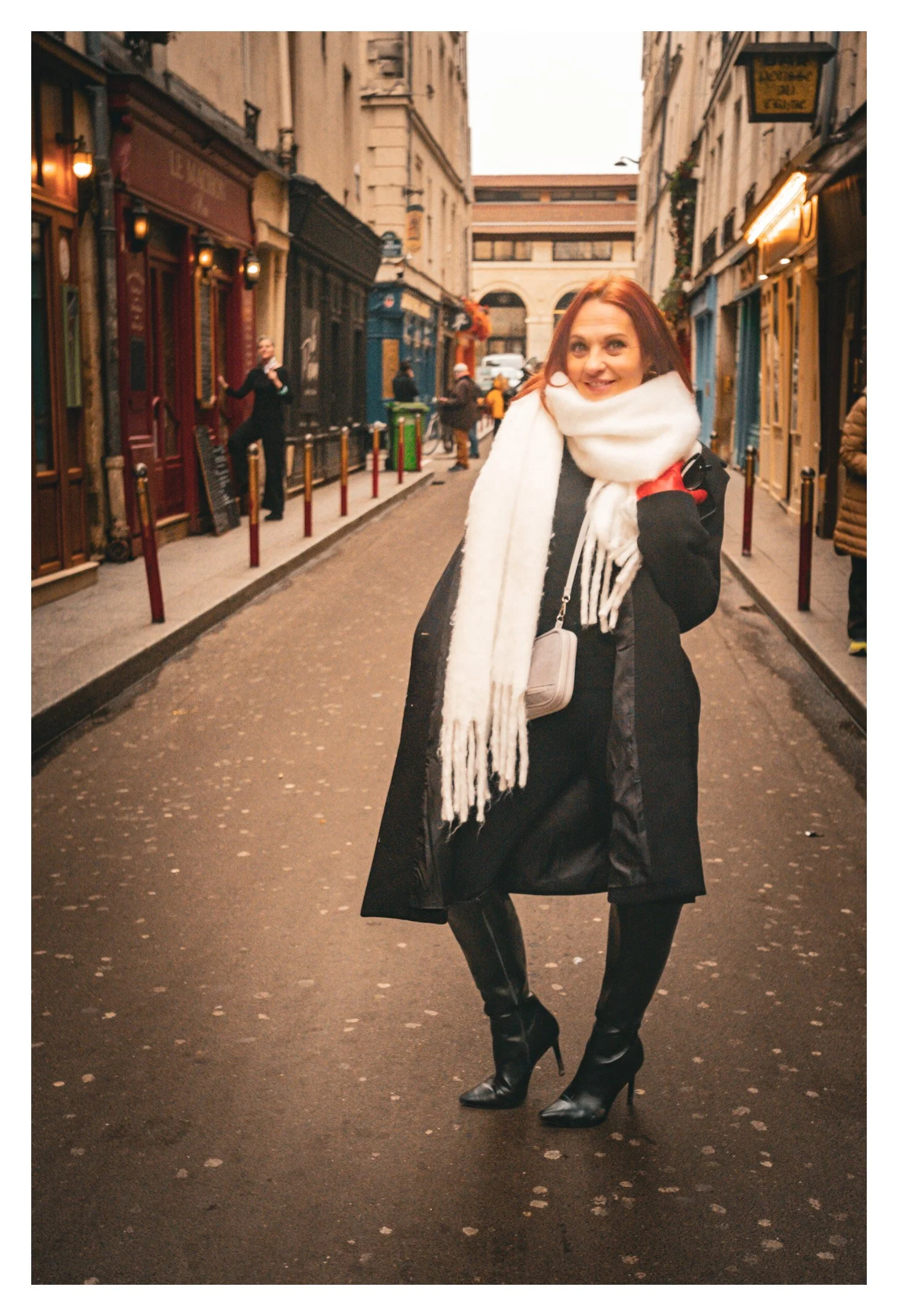 A woman with red hair smiling and posing on a narrow city street in front of colorful storefronts, wearing a black coat, large white scarf, and high heeled black boots.
