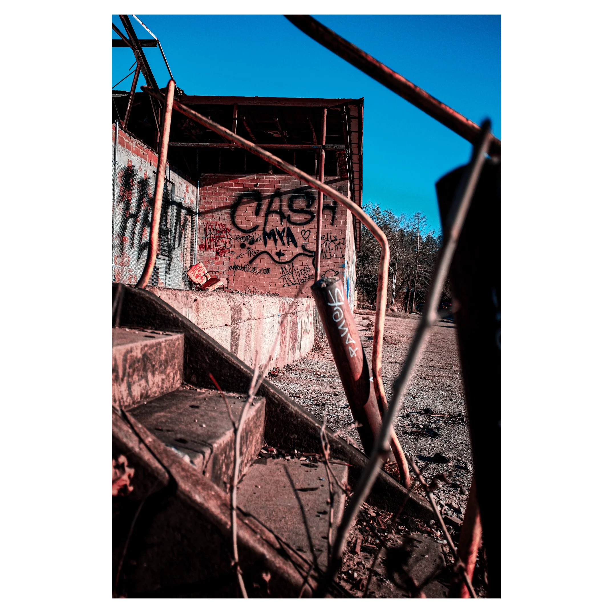 A rusted chain-link fence with graffiti on a brick building in the background, featuring the word 'CASH' in large black letters, under a clear blue sky.