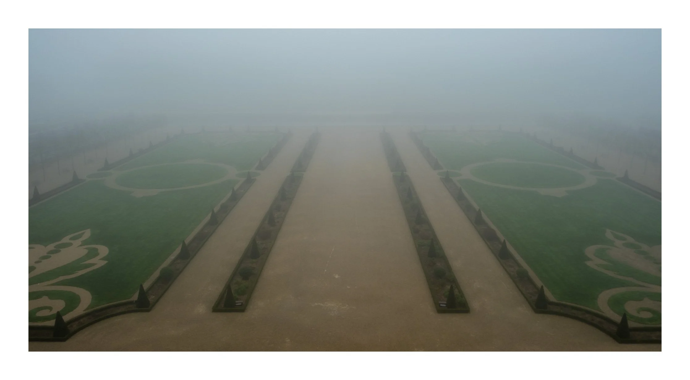 A foggy garden with symmetrical green lawn and pathways, surrounded by small bushes and pointed topiary structures.