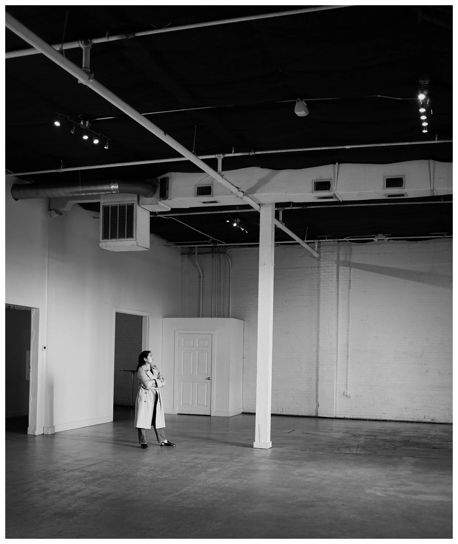 A woman in a trench coat standing in an empty, industrial-style room with white brick walls, wooden floor, and exposed ceiling pipes, looking towards the ceiling or lighting.