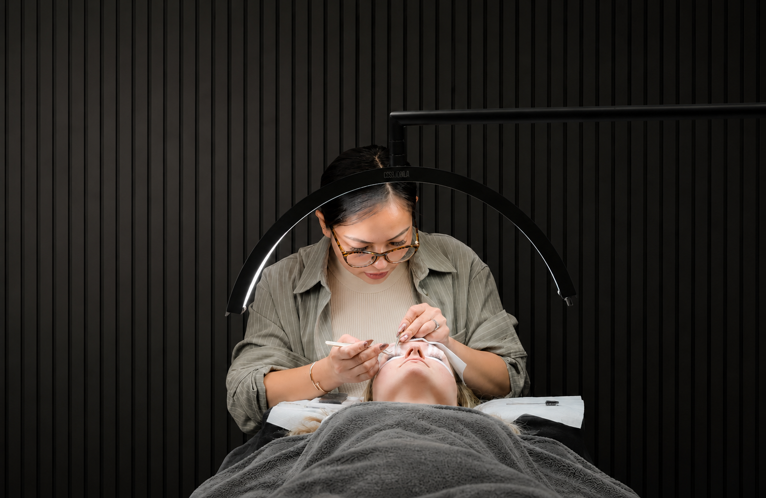 A woman performs a cosmetic procedure on a woman's eyes in a modern, dark clinic. The practitioner is wearing glasses and focusing on the patient's eyes, using tools under a circular overhead light.