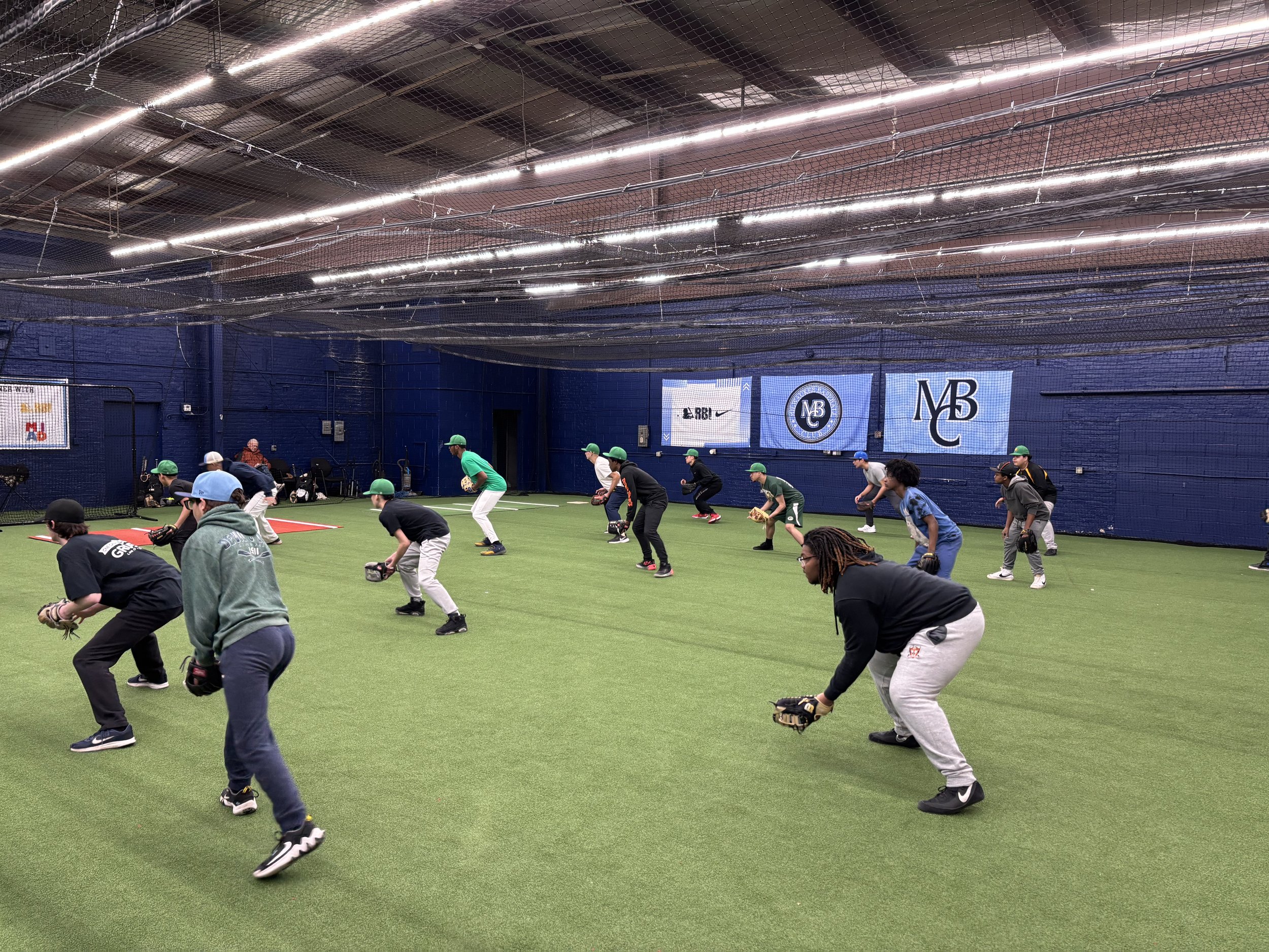Indoor baseball training session with young players practicing fielding on artificial turf, wearing caps and gloves, in a facility with banners on the wall.