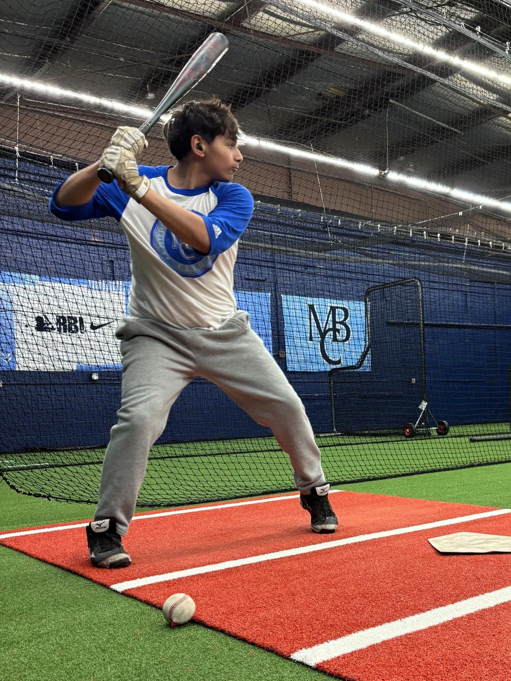 A man in a blue and white baseball shirt practicing batting in an indoor sports facility, holding a bat in a ready position on a red mat near a baseball and a home plate.