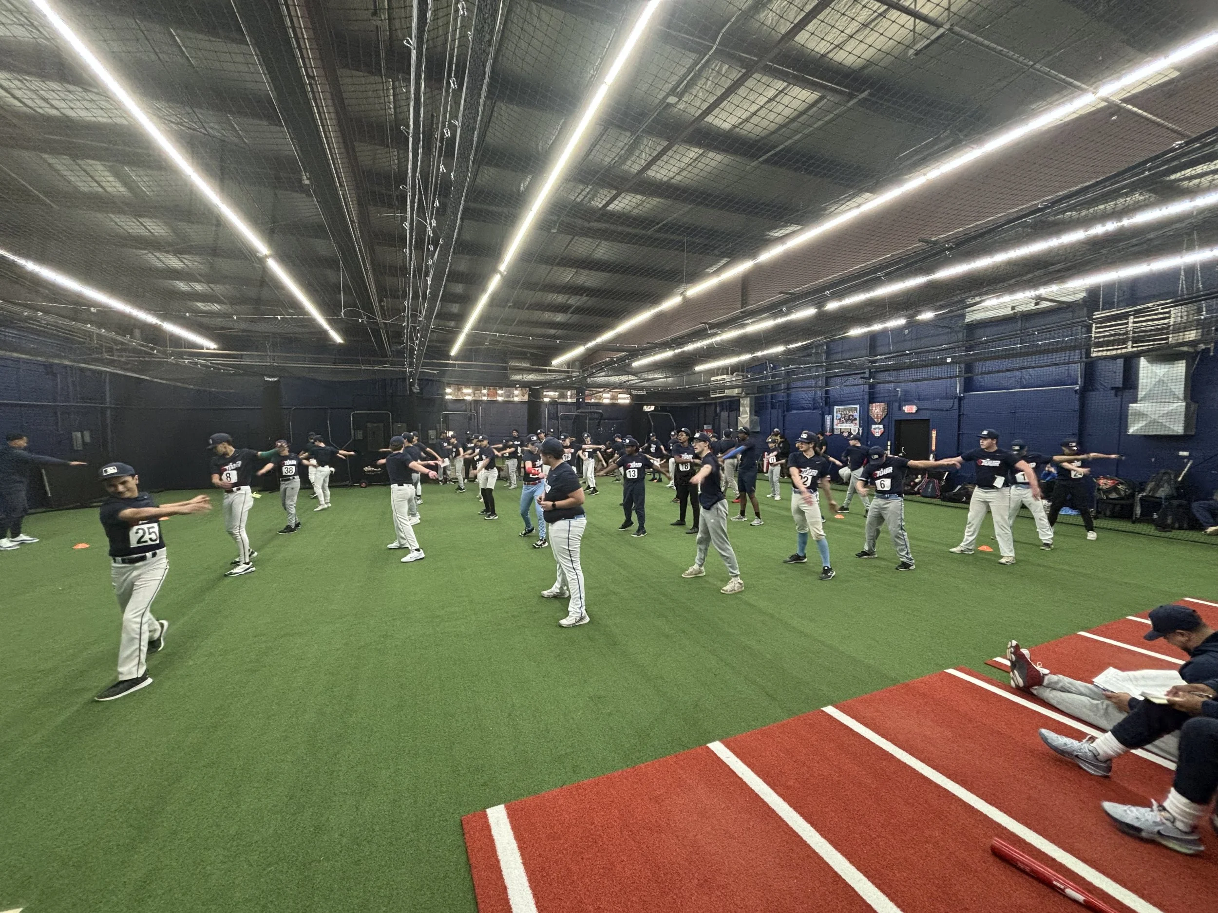 Indoor baseball training session with players warming up on artificial turf, wearing uniforms with numbers, in a well-lit facility.